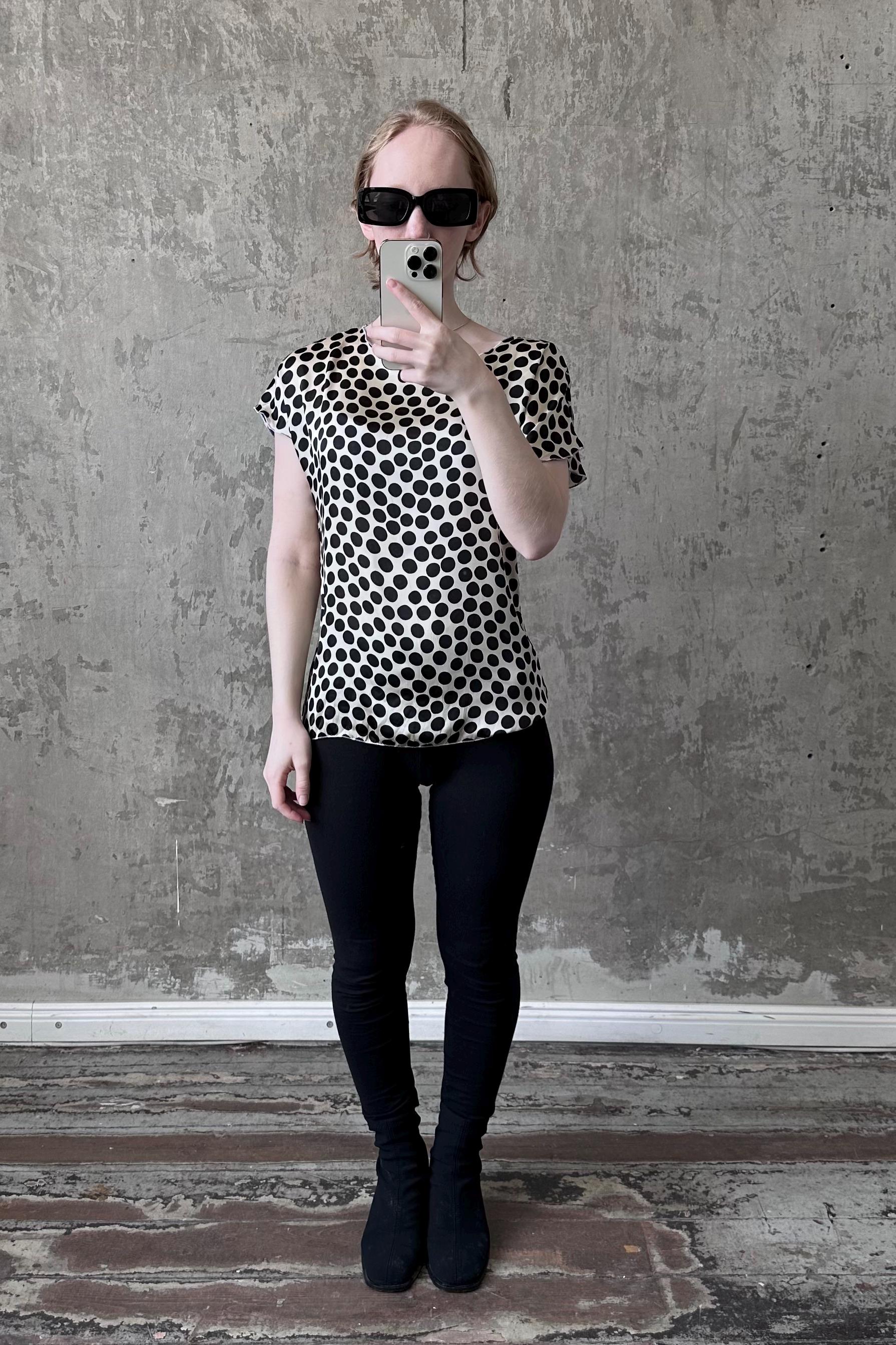 A woman wearing a black and white polka dot blouse and black leggings is taking a selfie with a smartphone against a concrete wall.