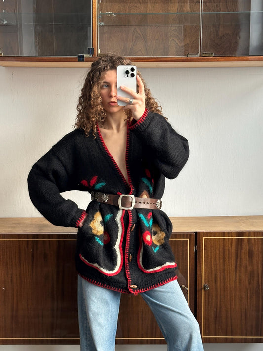 A woman with curly hair is taking a selfie with a smartphone, wearing a black sweater with colorful floral patterns and a brown belt.