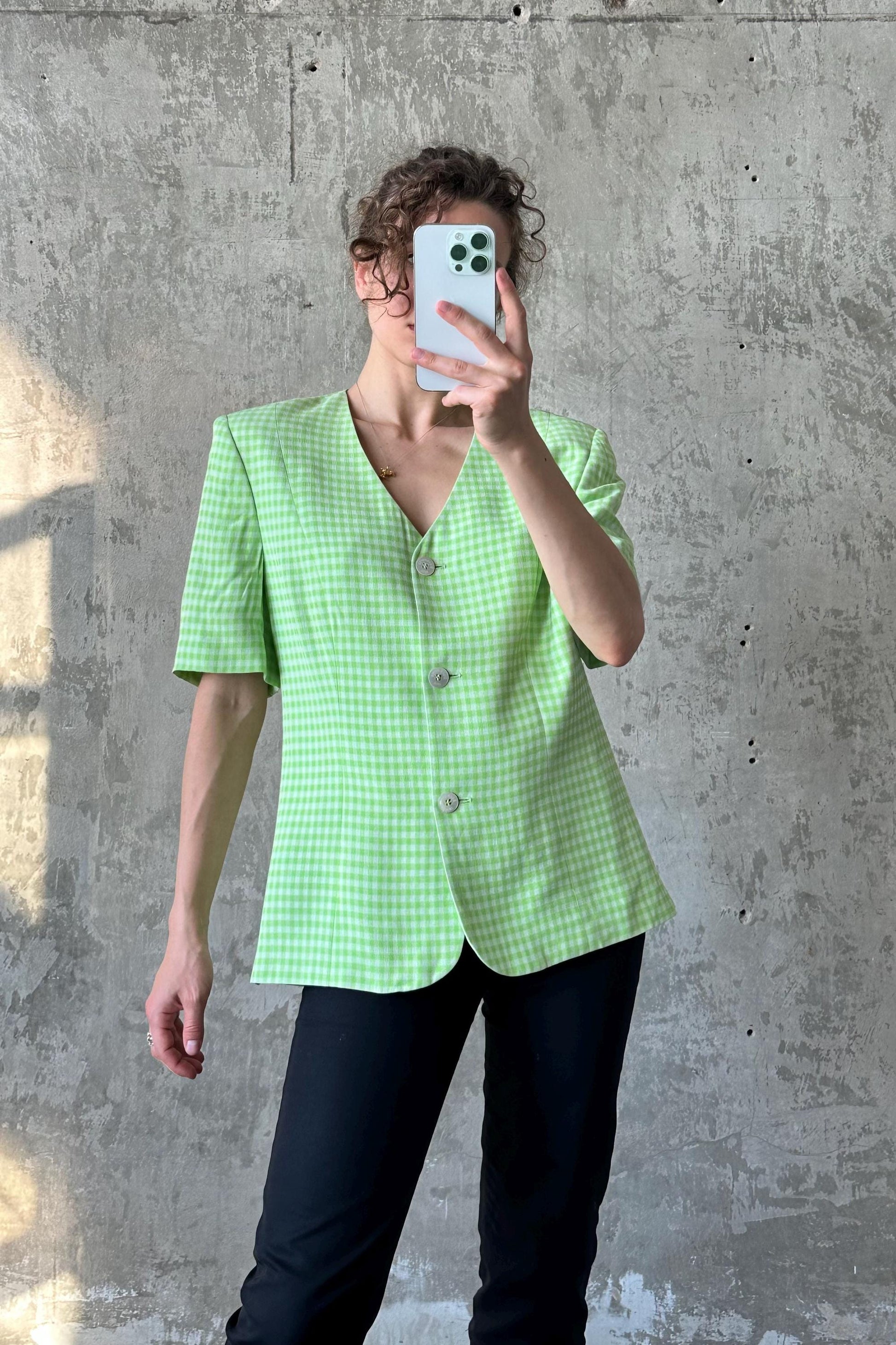 A person is taking a selfie with a white phone, wearing a green and white checkered blazer and black pants.