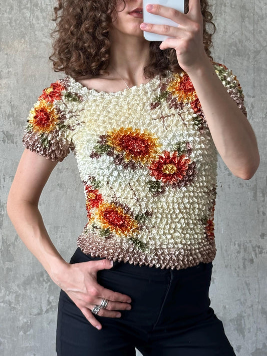 A woman with curly hair is taking a selfie with a white phone, wearing a floral print top and black pants.