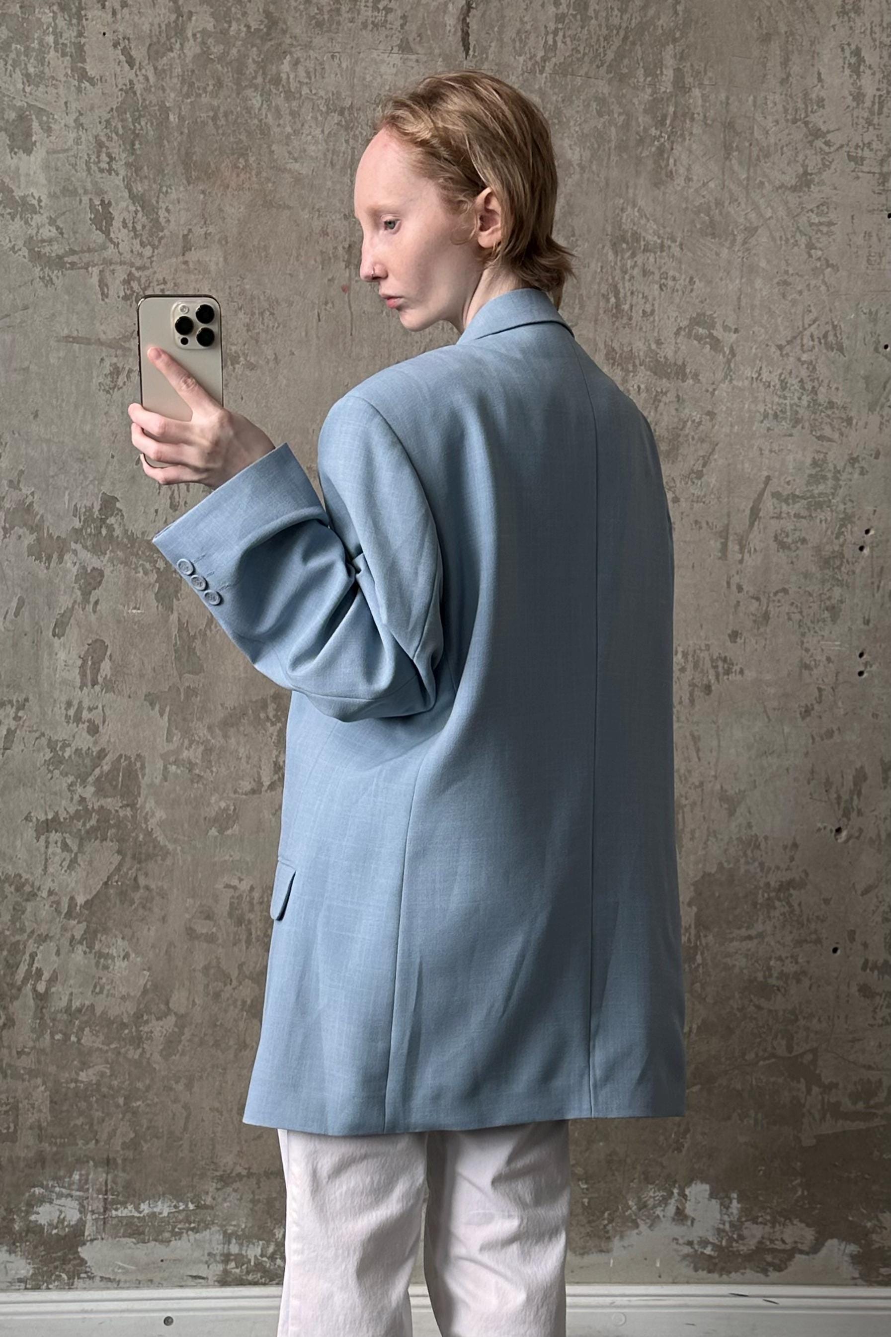 A person in a light blue blazer is holding a smartphone and looking at it, standing against a textured concrete wall.