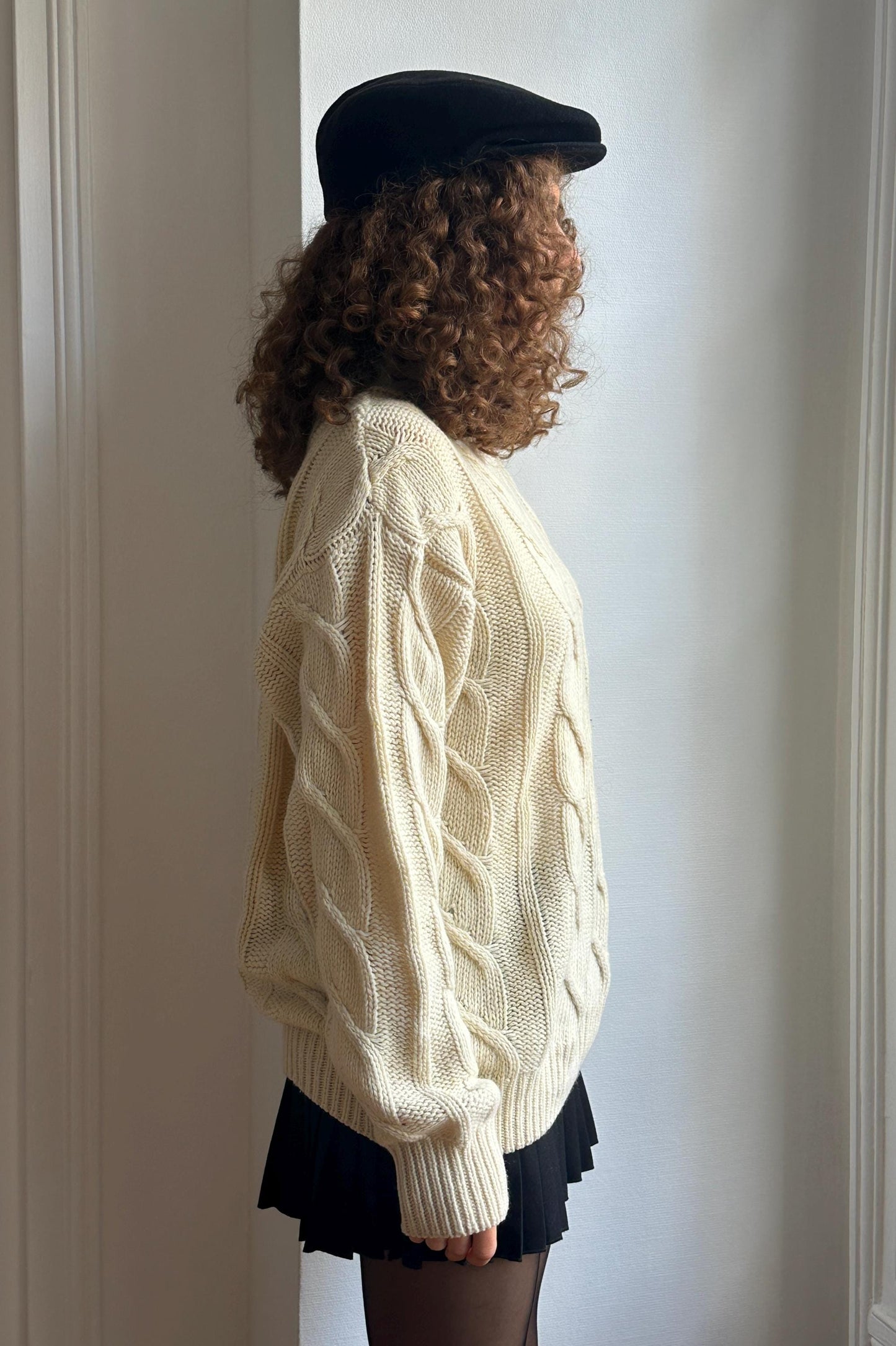 A person with curly hair wearing a white sweater and a black hat stands in front of a white wall.