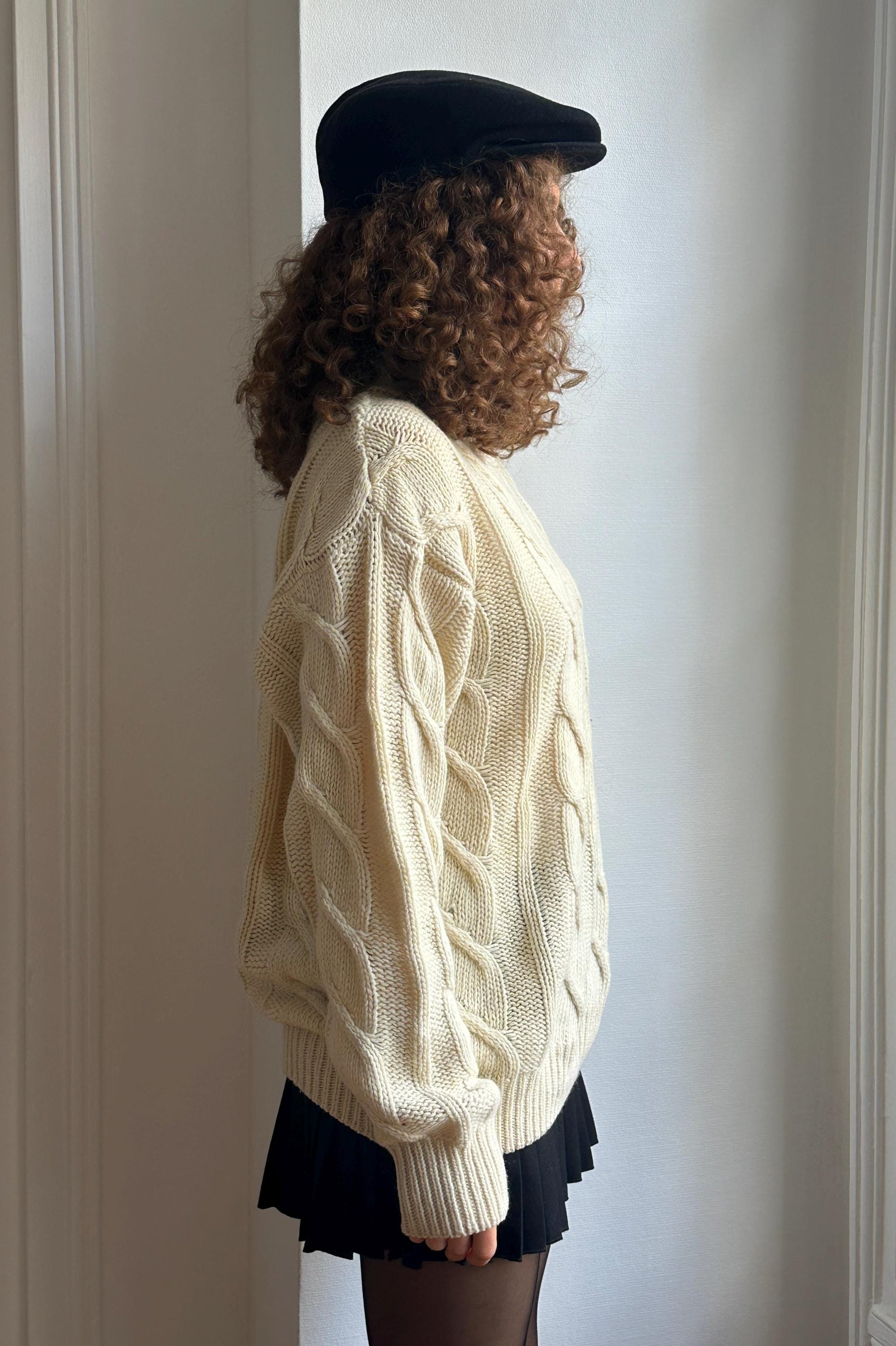 A person with curly hair wearing a white sweater and a black hat stands in front of a white wall.