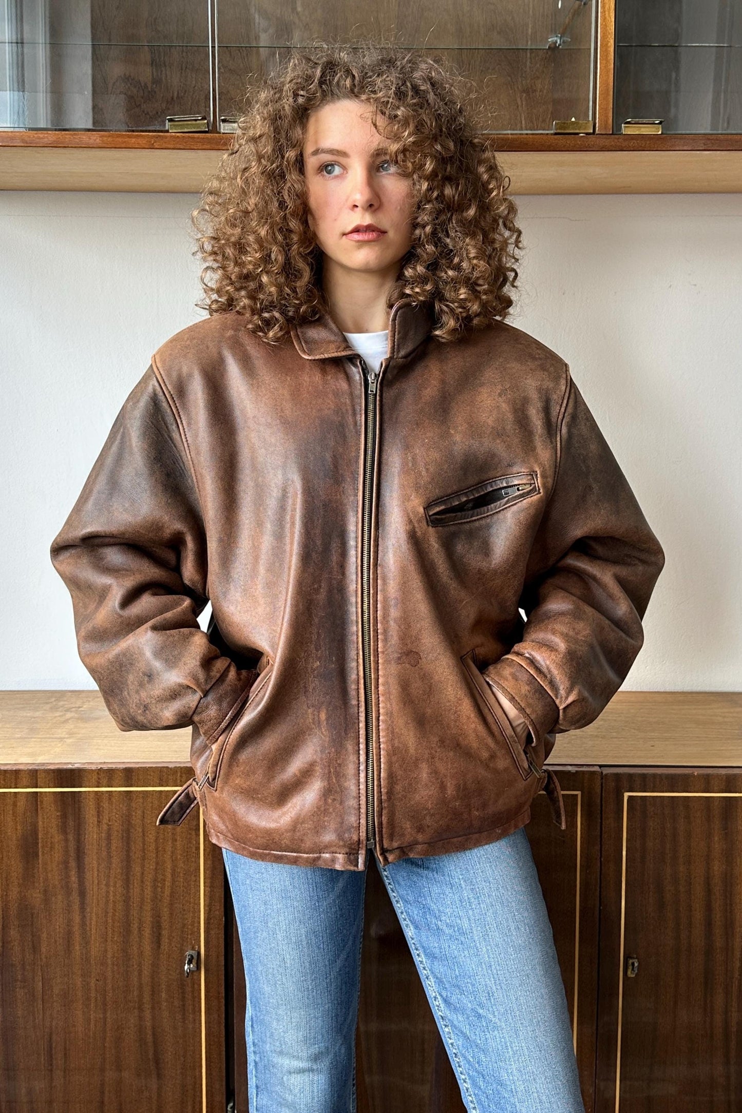 A woman with curly hair wearing a brown leather jacket stands in front of a wooden cabinet.