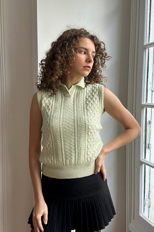A young woman with curly hair stands in front of a window, wearing a sleeveless white sweater and a black skirt.