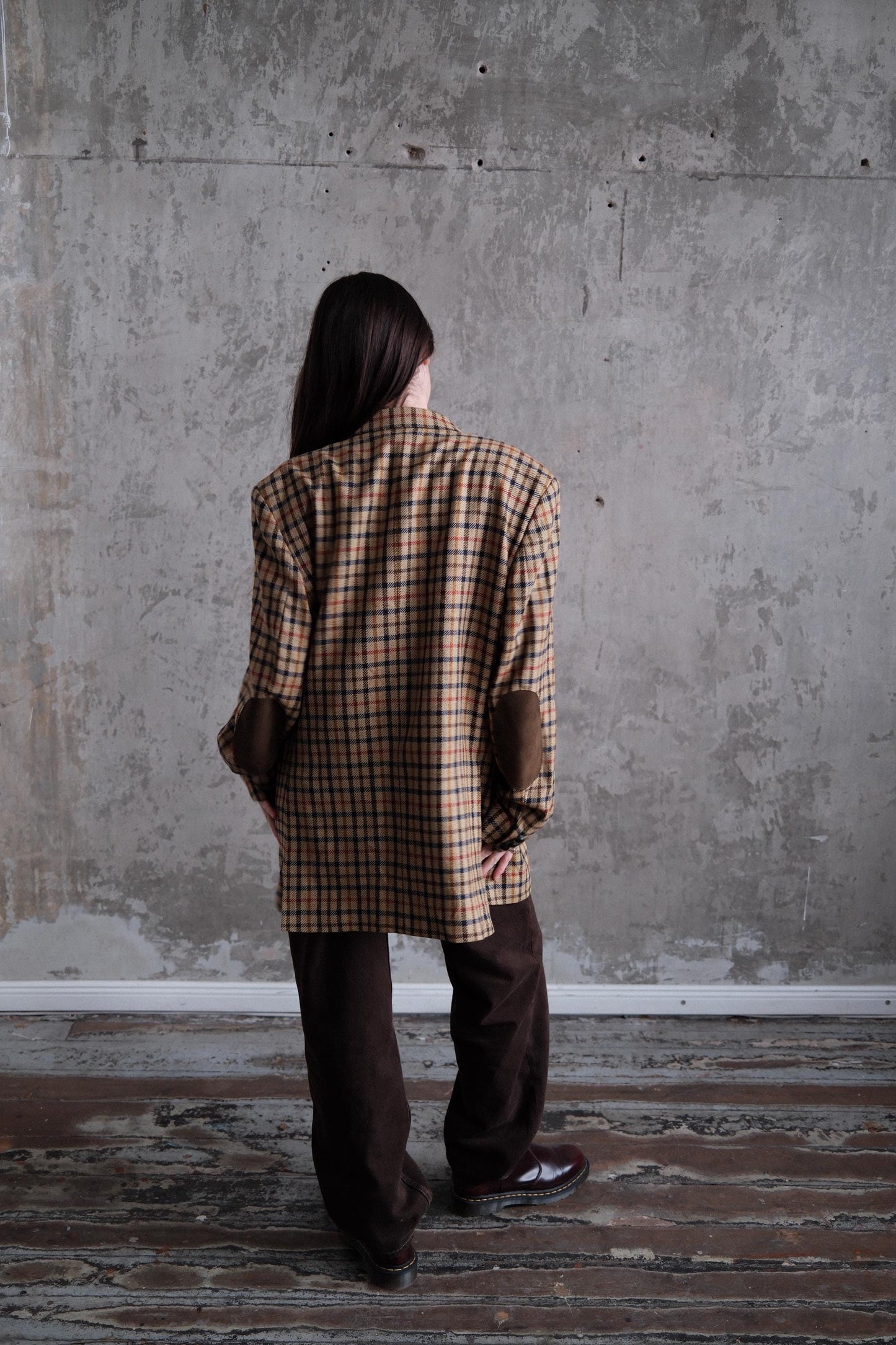 a person standing in a room with a concrete wall and a wooden floor. The person is wearing a plaid jacket and brown pants.