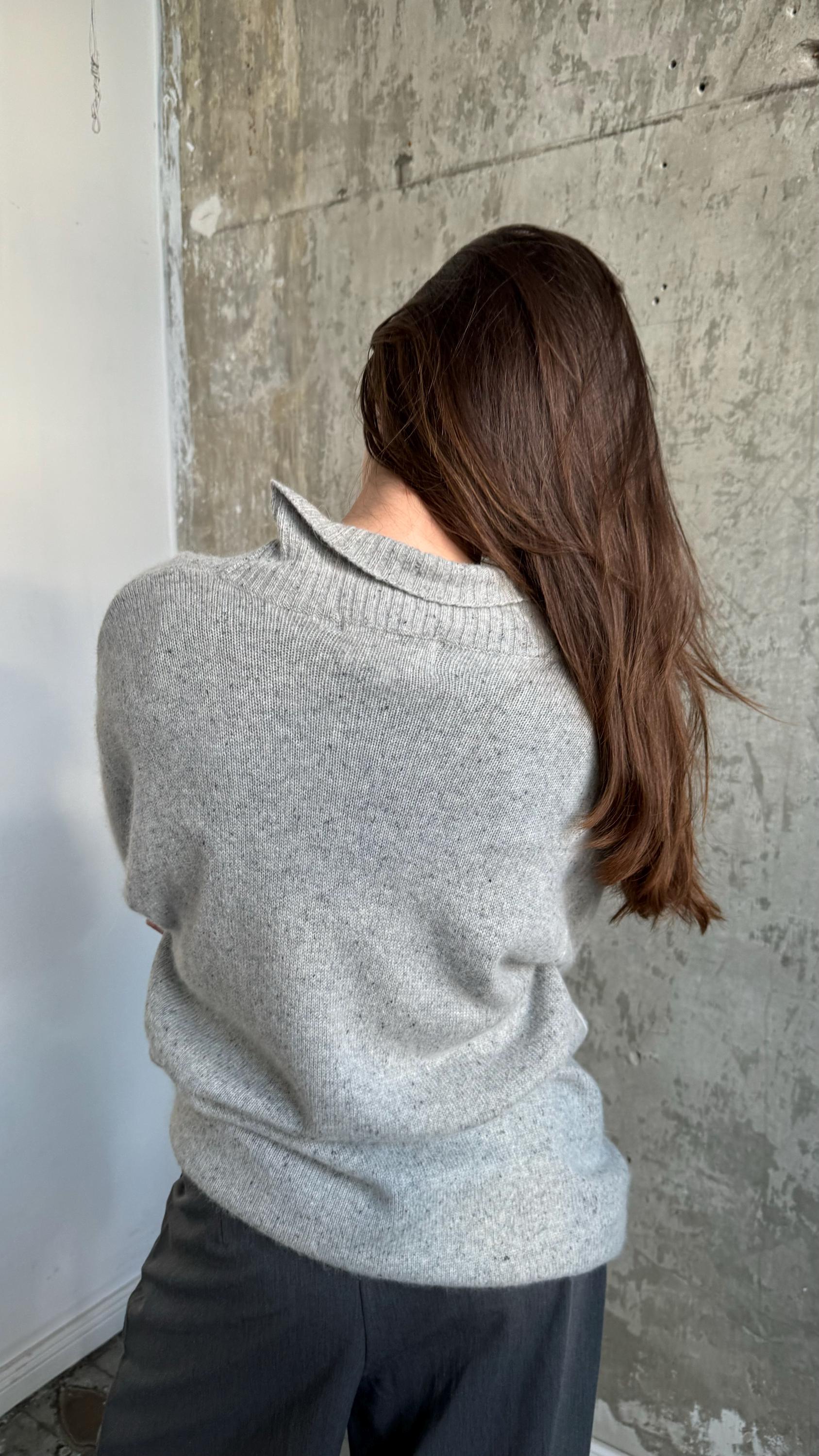a person with long brown hair wearing a gray sweater, standing in front of a concrete wall.
