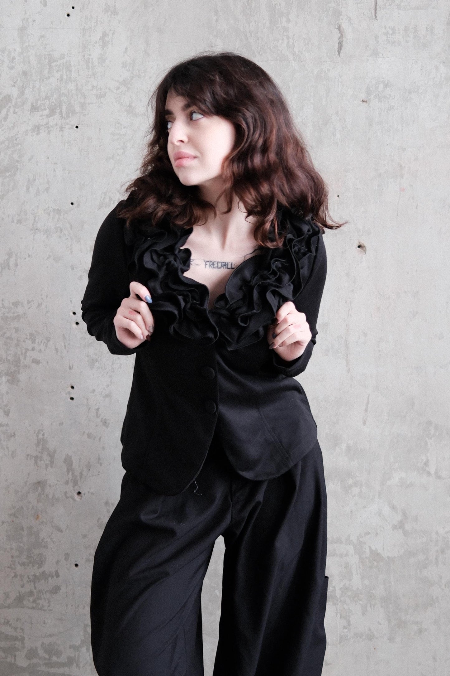 A woman with long, dark hair is wearing a black outfit and posing in a relaxed, contemplative manner against a plain background.