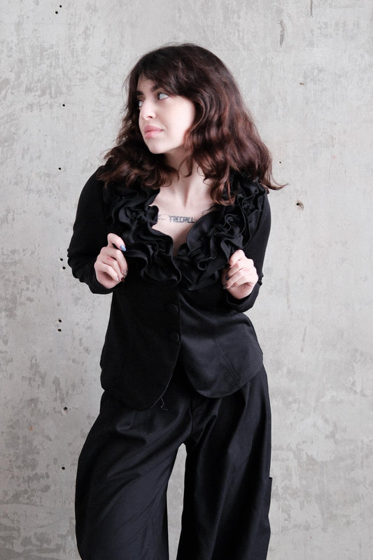 A woman with long, dark hair is wearing a black outfit and posing in a relaxed, contemplative manner against a plain background.