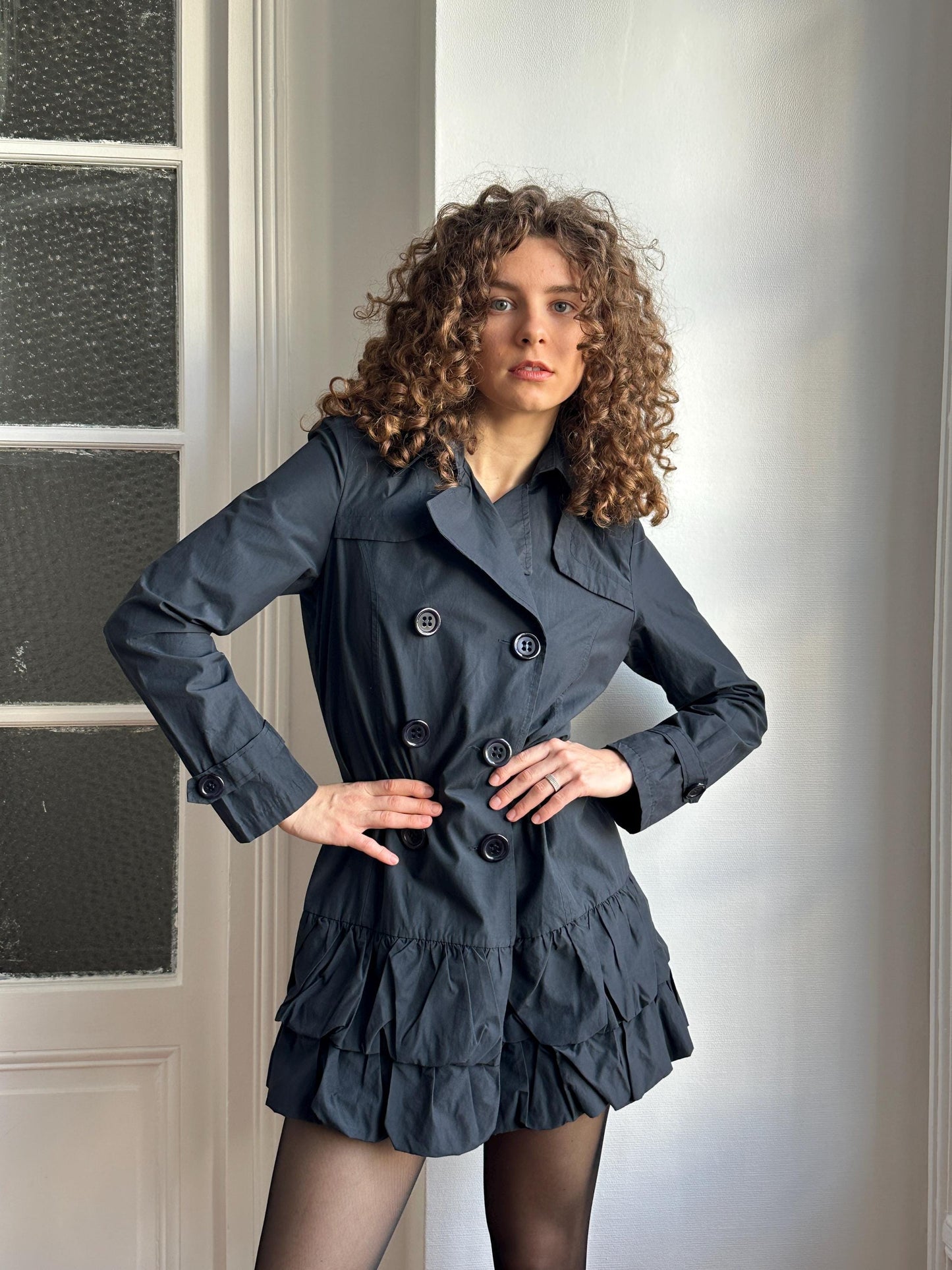 A woman with curly hair wearing a black coat and skirt is posing in a relaxed manner, with her hands on her hips.