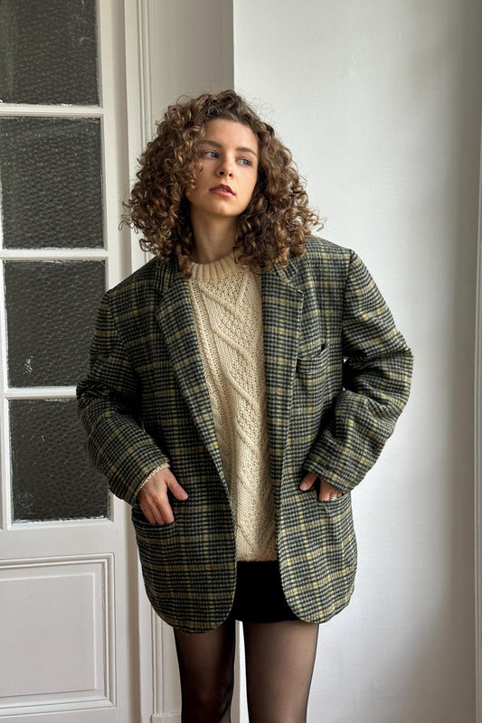 A young woman with curly hair stands in front of a white door, wearing a plaid jacket, a white sweater, and black shorts.