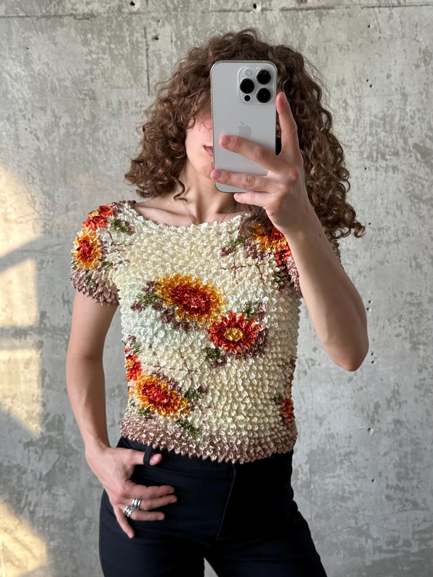 A person with curly hair is taking a selfie with a white iPhone, wearing a floral print top and black pants.