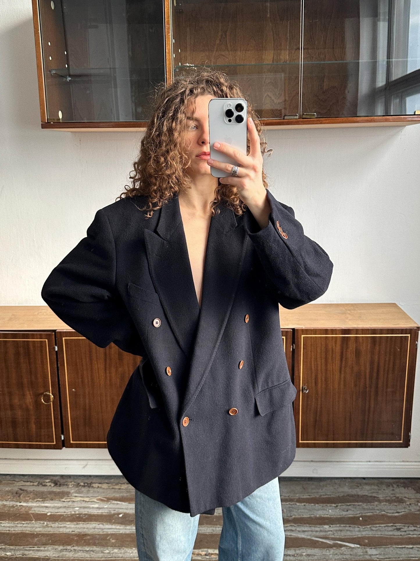 A person with curly hair is taking a selfie with a smartphone, wearing a black blazer and standing in front of a wooden cabinet.