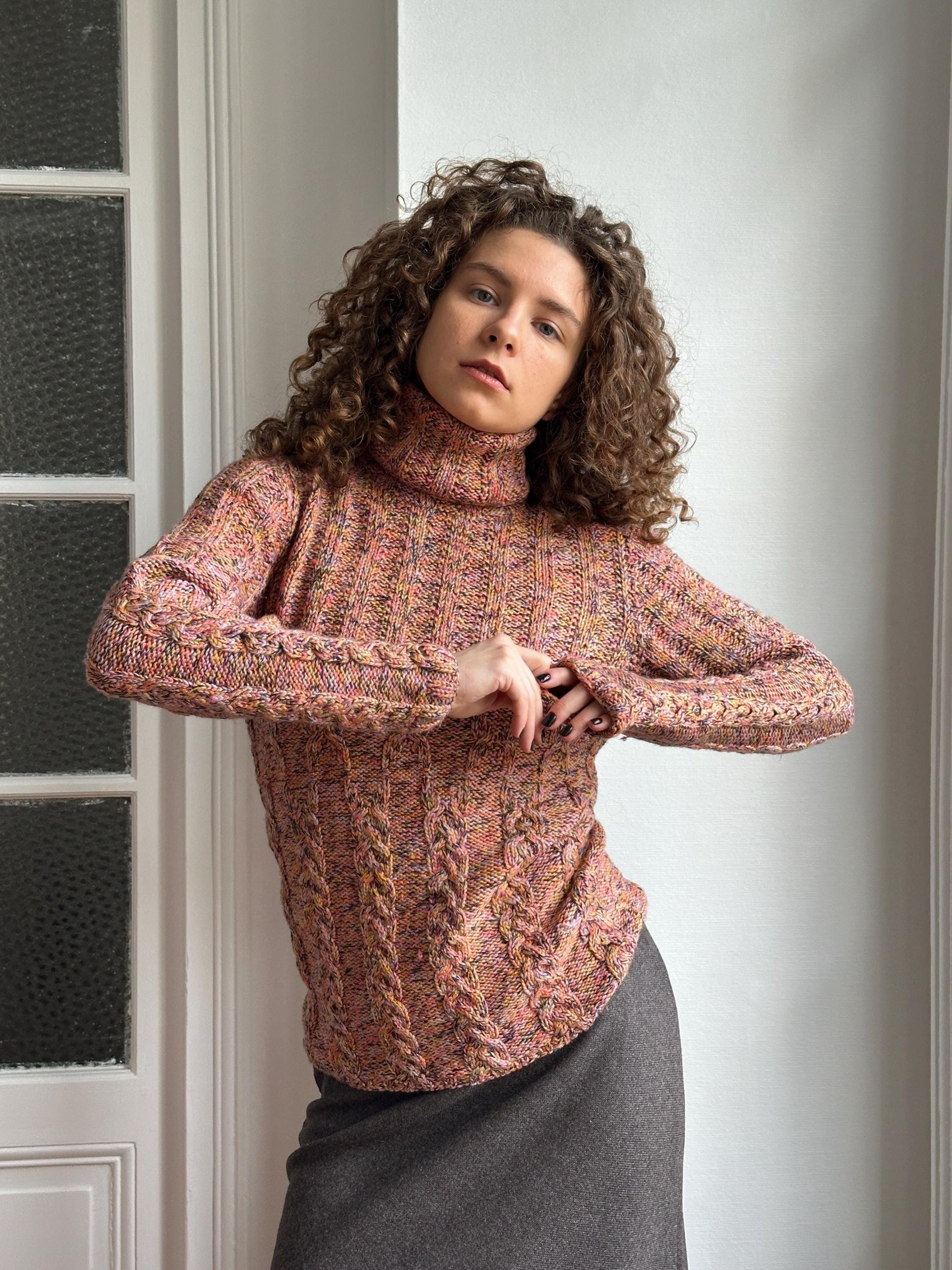 A woman with curly hair wearing a colorful, patterned sweater is posing in a relaxed, leaning position.
