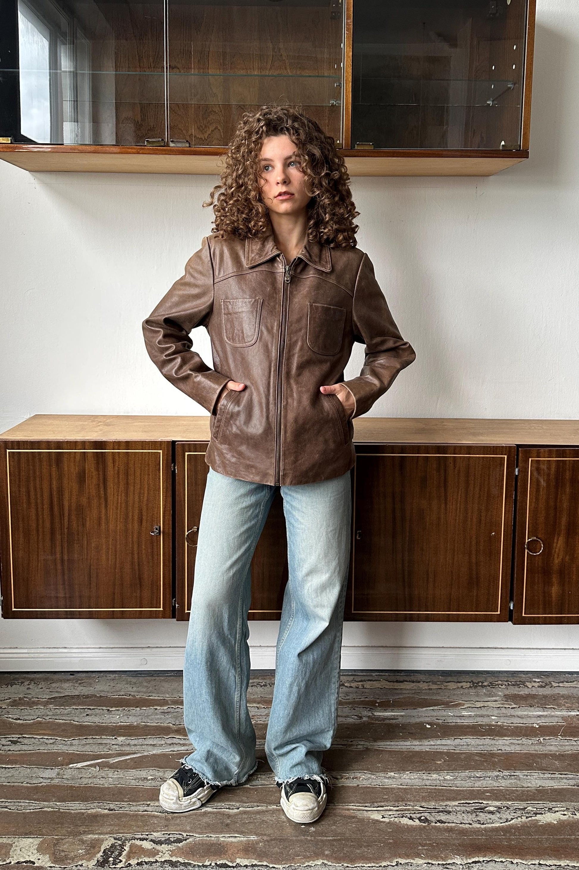 A woman with curly hair wearing a brown leather jacket and blue jeans stands in a room with wooden cabinets.