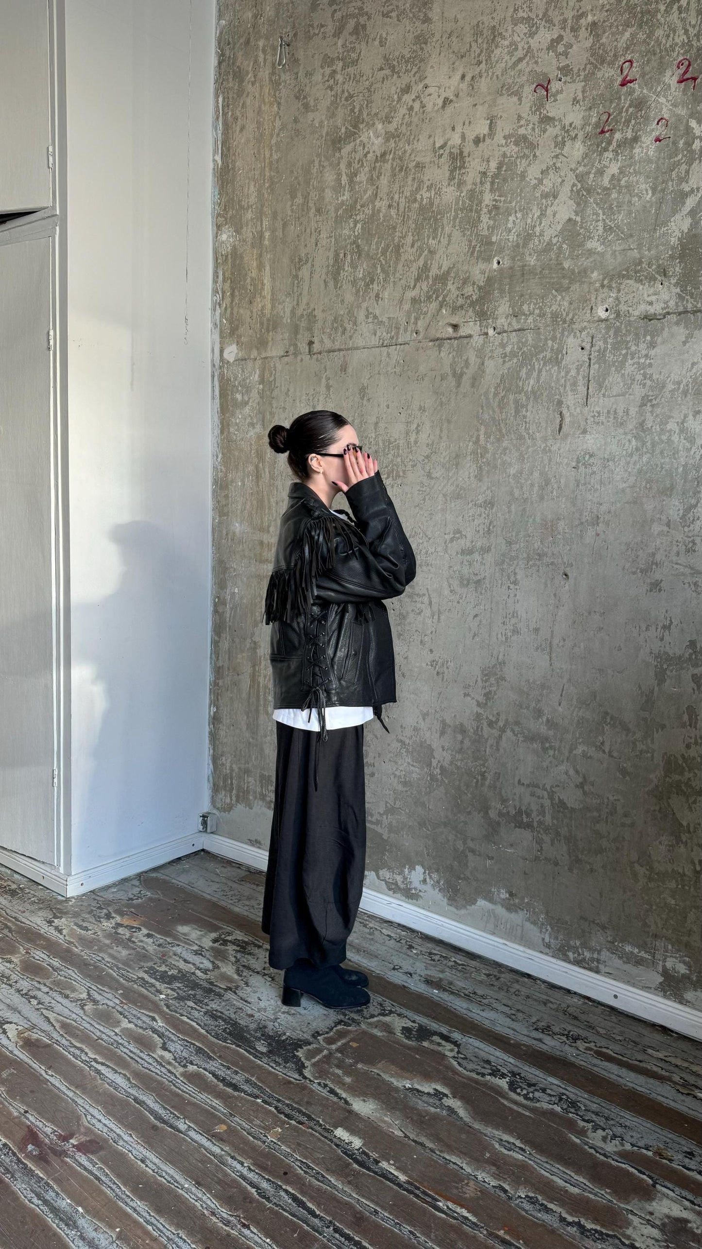 A person wearing a black leather jacket and sunglasses stands in a room with a concrete wall and wooden floor.