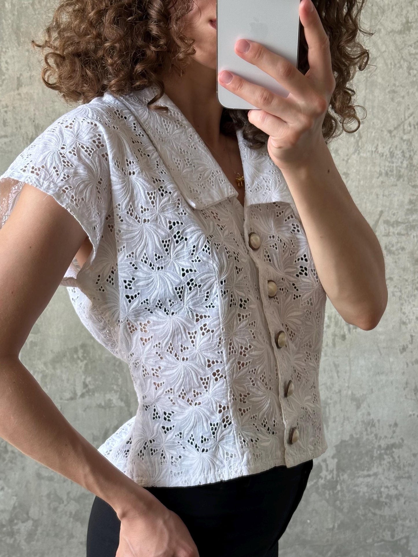A woman with curly hair is taking a selfie with a white lace blouse featuring a floral pattern and buttons.