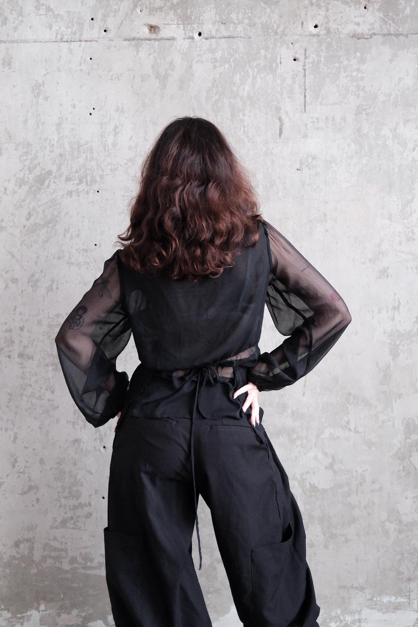 A person with long, curly hair wearing a black outfit stands against a plain concrete wall.