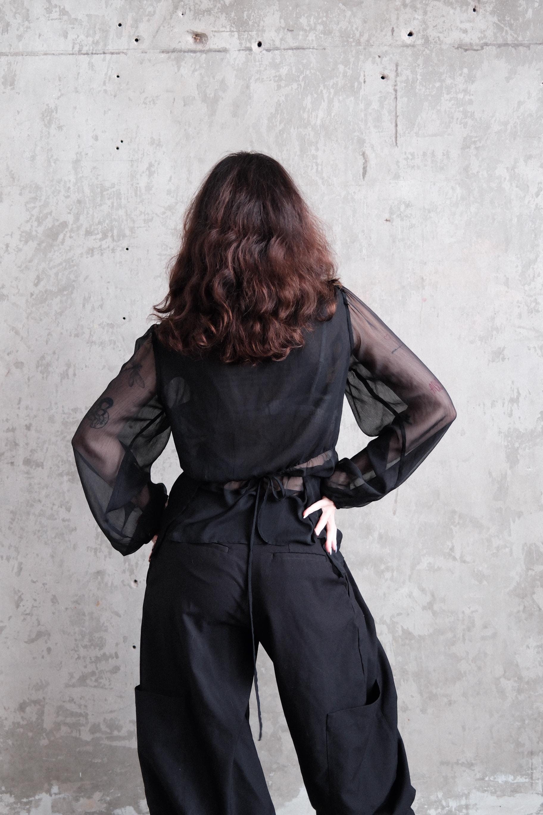 A person with long, curly hair wearing a black outfit stands against a plain concrete wall.