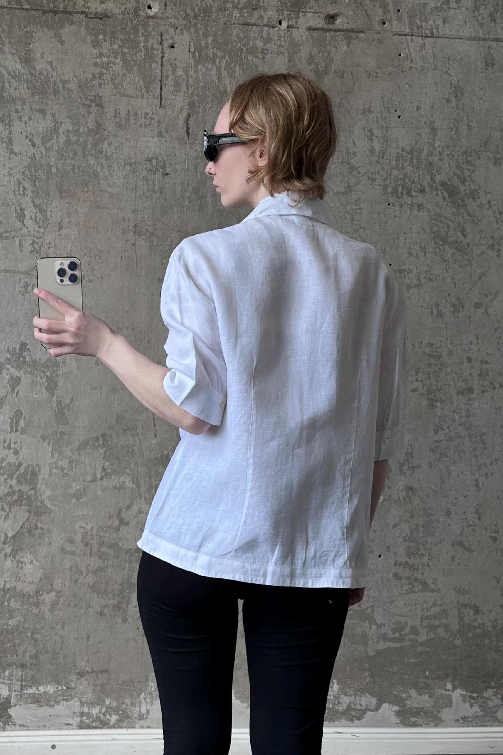 A person wearing a white shirt and sunglasses is holding a phone in front of a concrete wall.