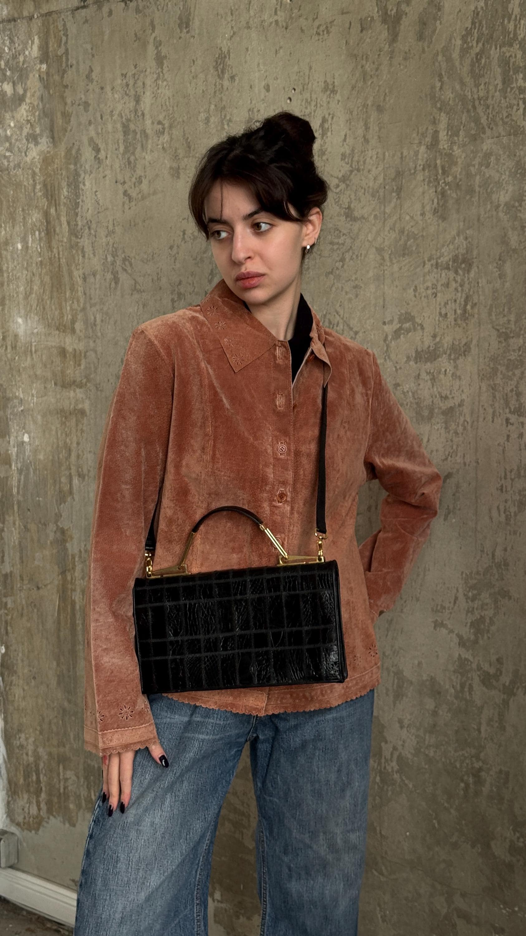 A young woman wearing a brown jacket and jeans stands against a concrete wall, holding a black handbag.