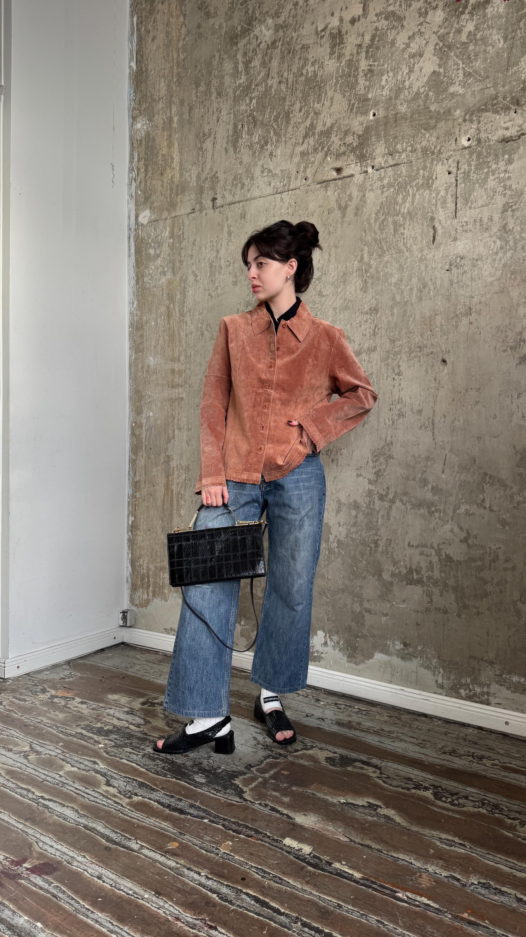 A woman wearing a brown jacket, blue jeans, and black sandals stands in a room with a concrete wall. She is holding a black bag in her hand.