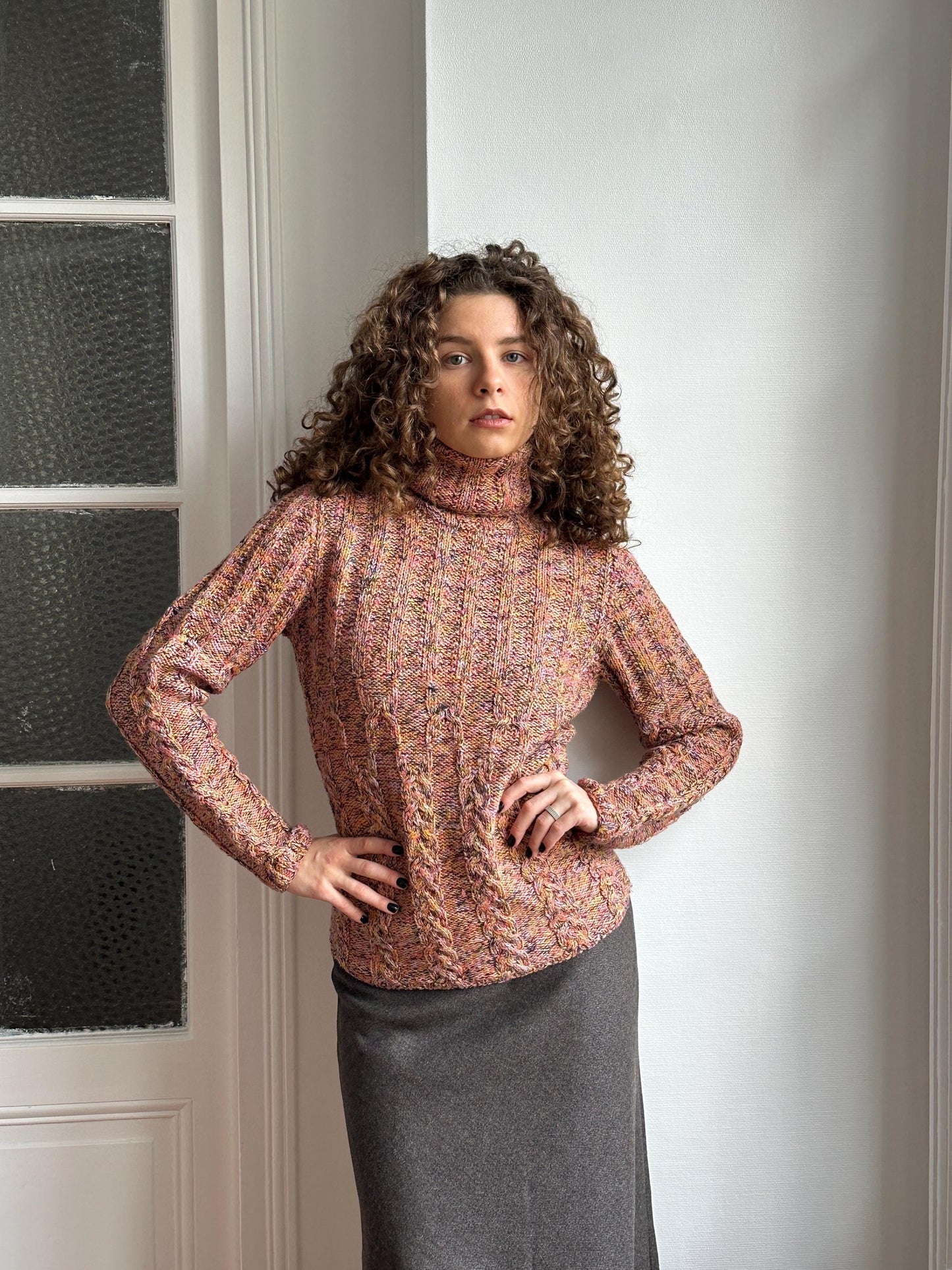 A woman with curly hair is posing in a room with a white wall and a window. She is wearing a colorful sweater and a gray skirt.