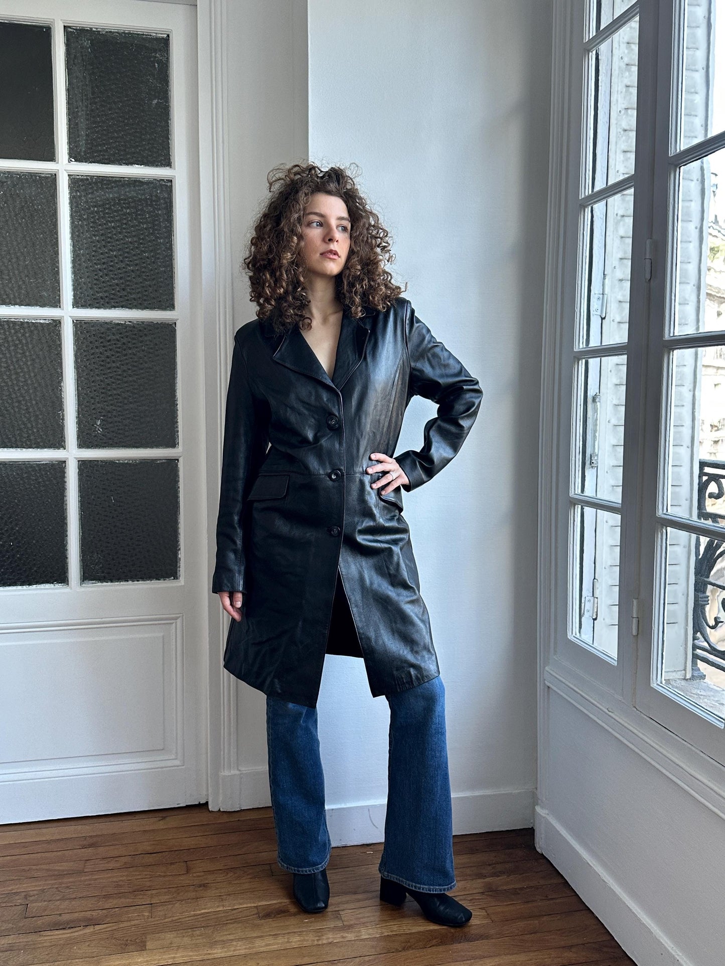 A woman in a black leather coat stands in a room with large windows, looking to the side with her hands on her hips.