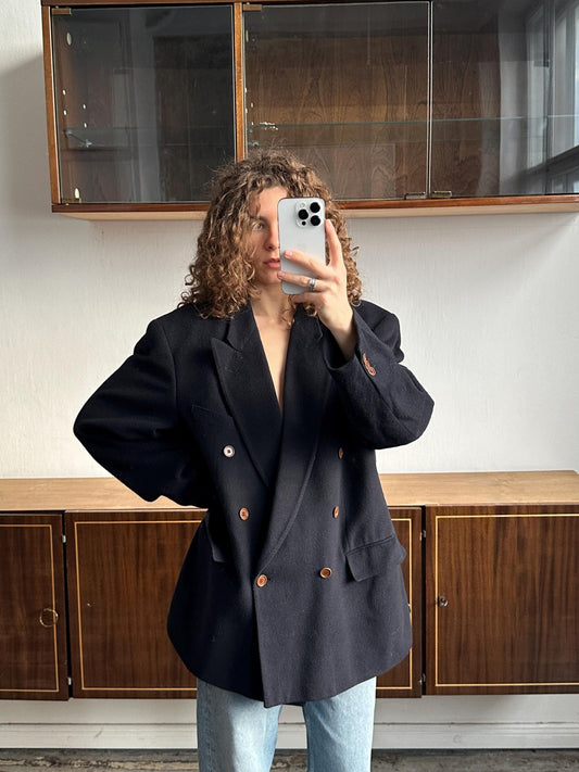 A person with curly hair is taking a selfie with a smartphone, wearing a black blazer and standing in front of a wooden cabinet.
