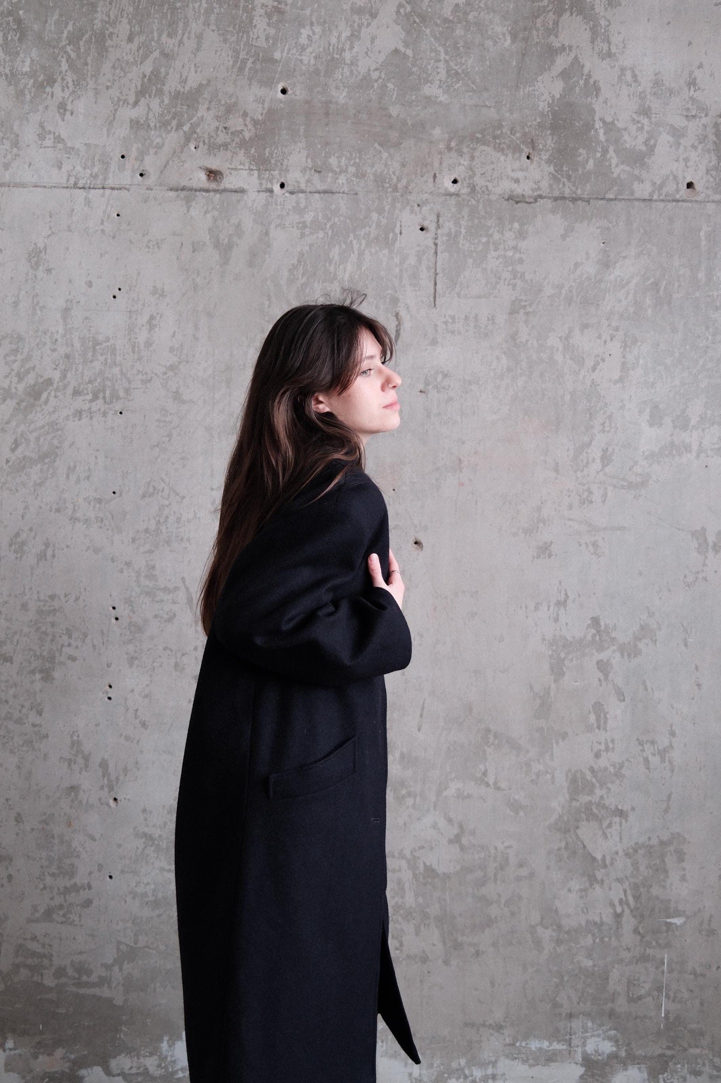 A woman in a black coat is standing on a concrete floor, looking down at the ground.