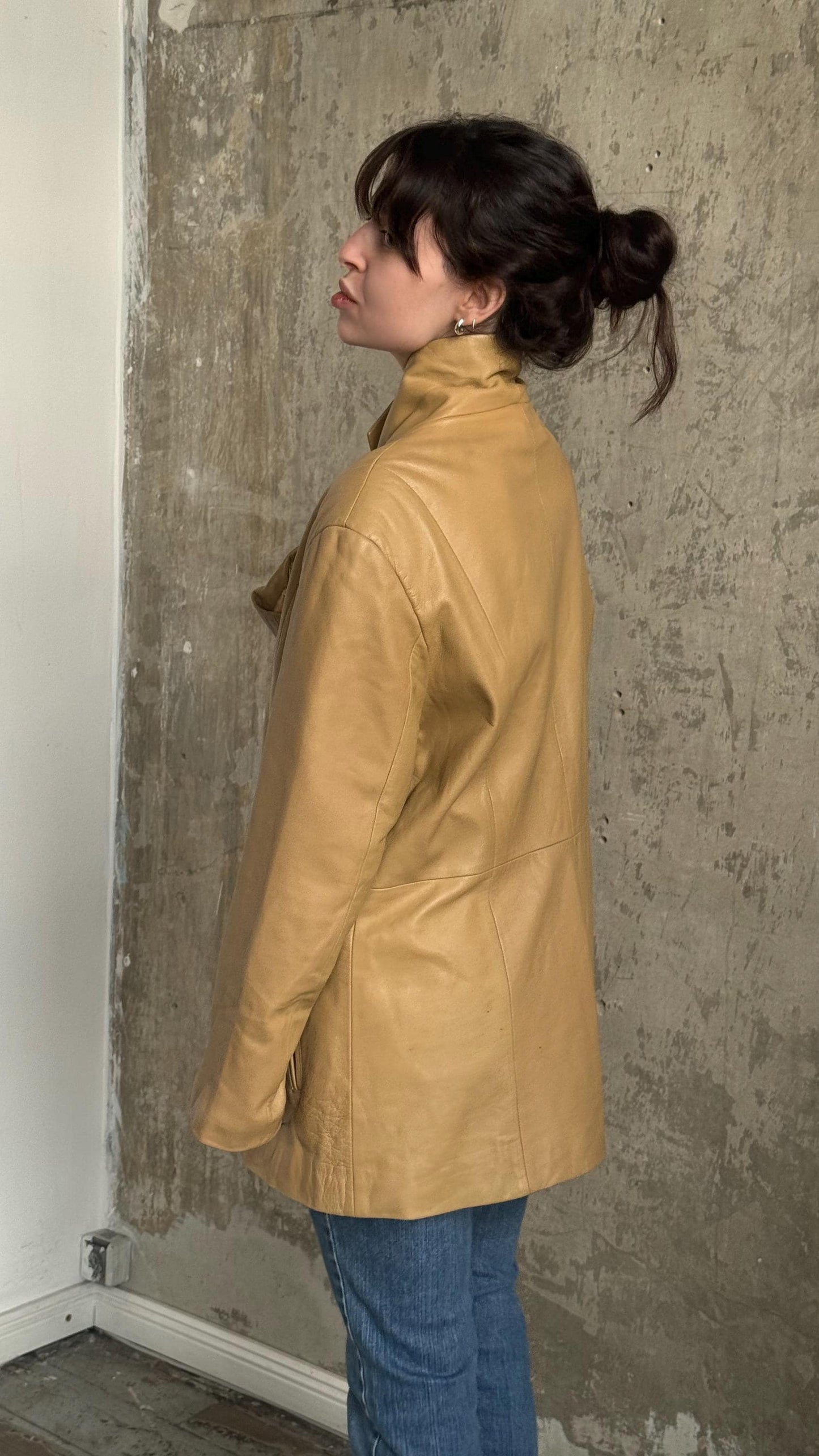 A woman wearing a tan leather jacket stands in front of a concrete wall, her back turned to the camera.