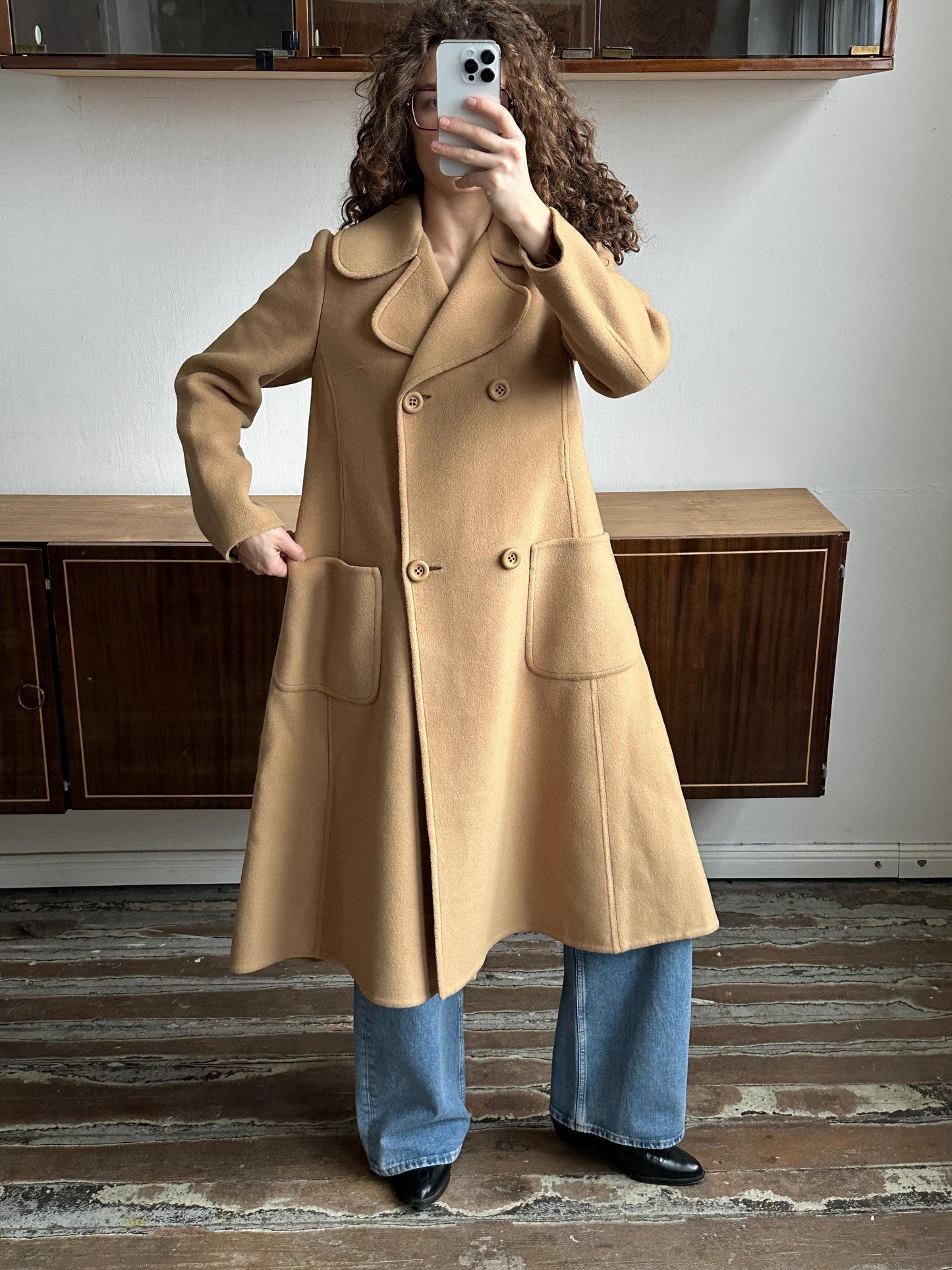 May include: A tan double-breasted coat with large front pockets and a flared skirt. The coat has a wide collar and is buttoned up. The person is wearing blue jeans and black shoes. The coat is a vintage style.