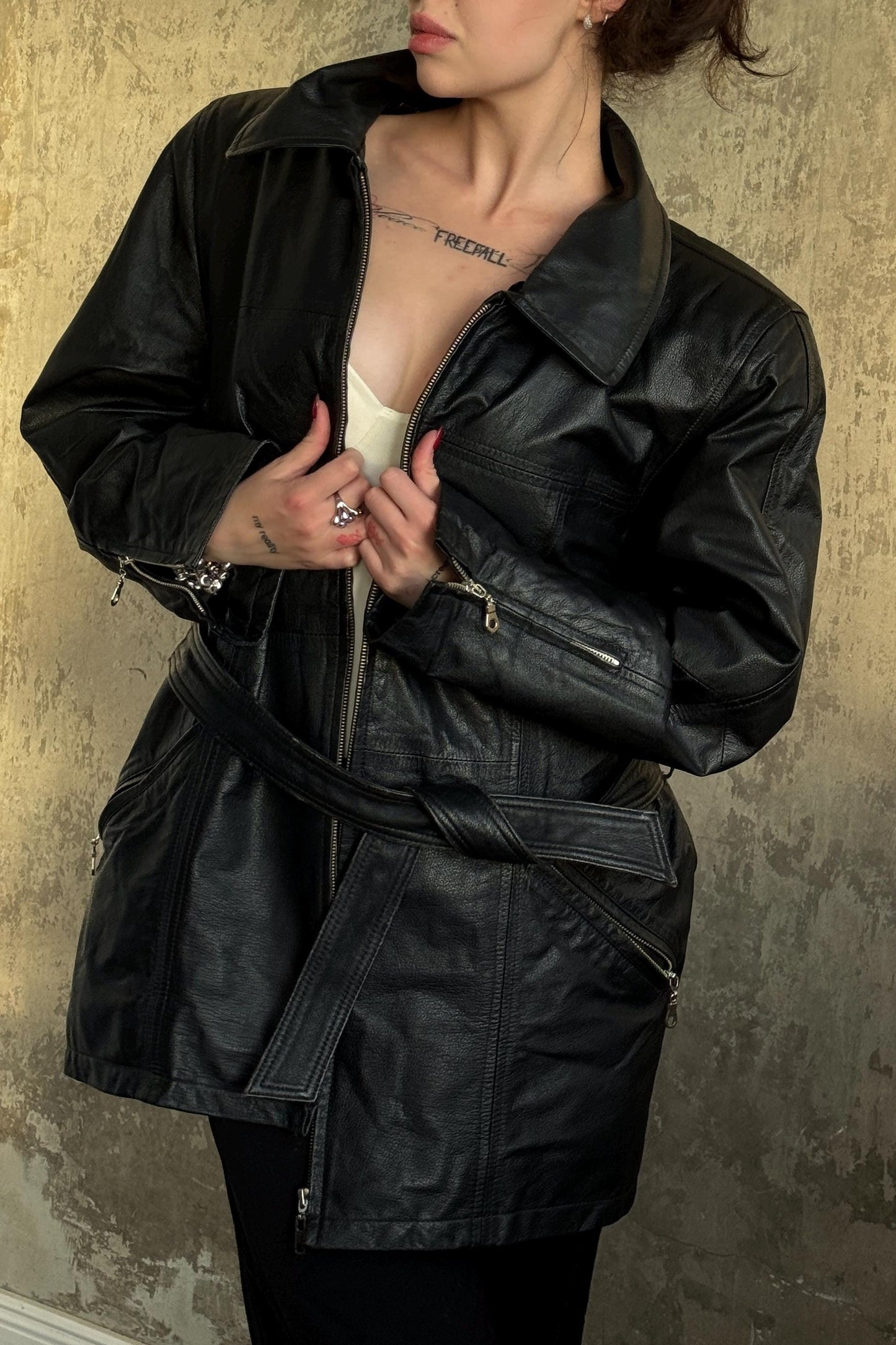 A woman wearing a black leather jacket stands against a textured wall.