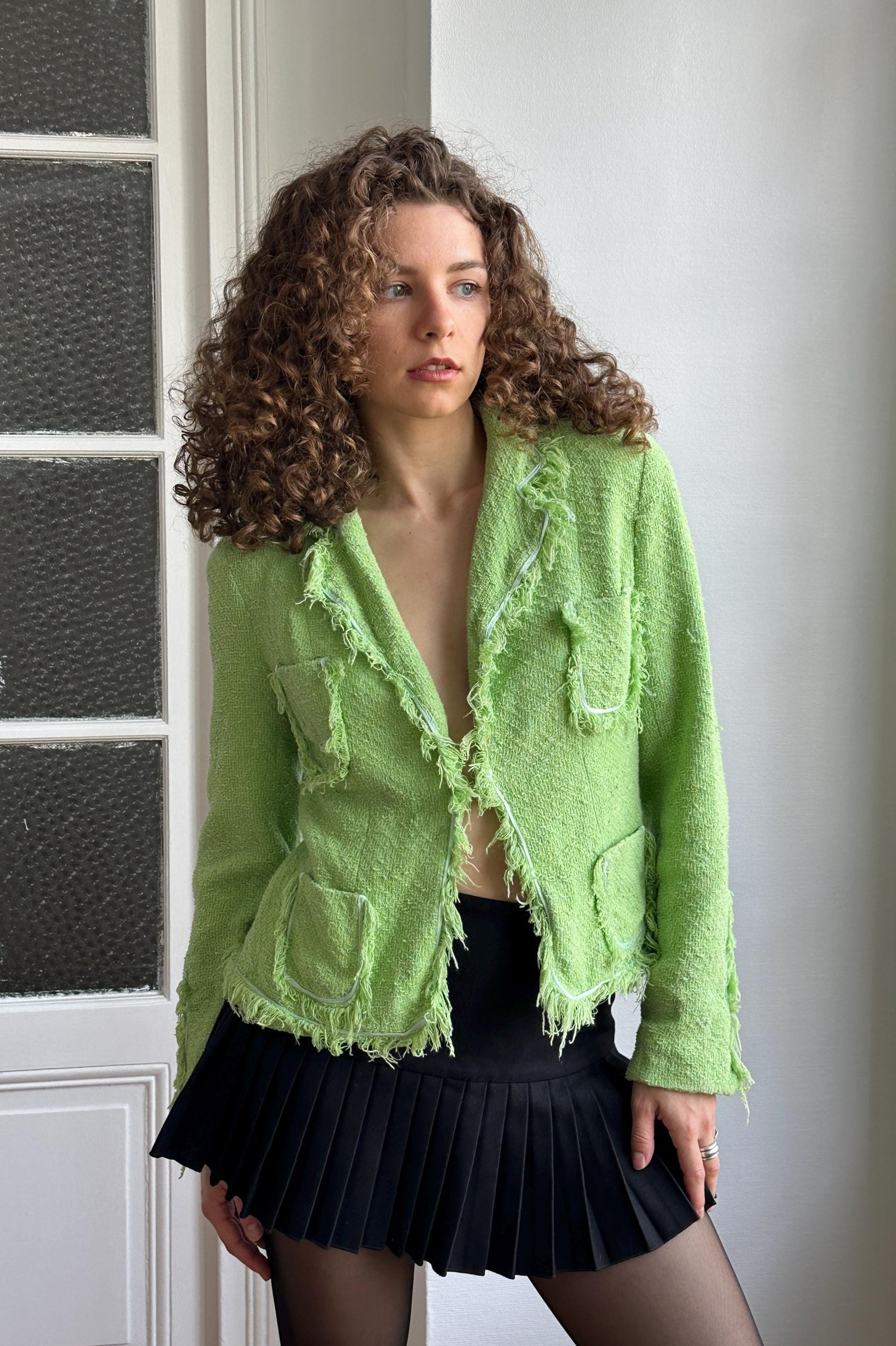 A woman with curly hair wearing a lime green jacket and a black pleated skirt stands in front of a white door.