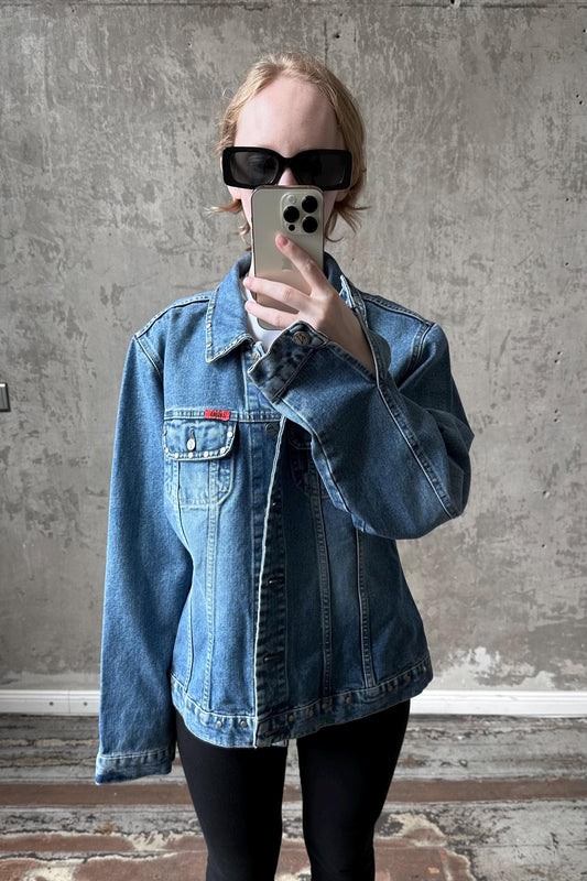 A person wearing a denim jacket and sunglasses is taking a selfie with a smartphone.
