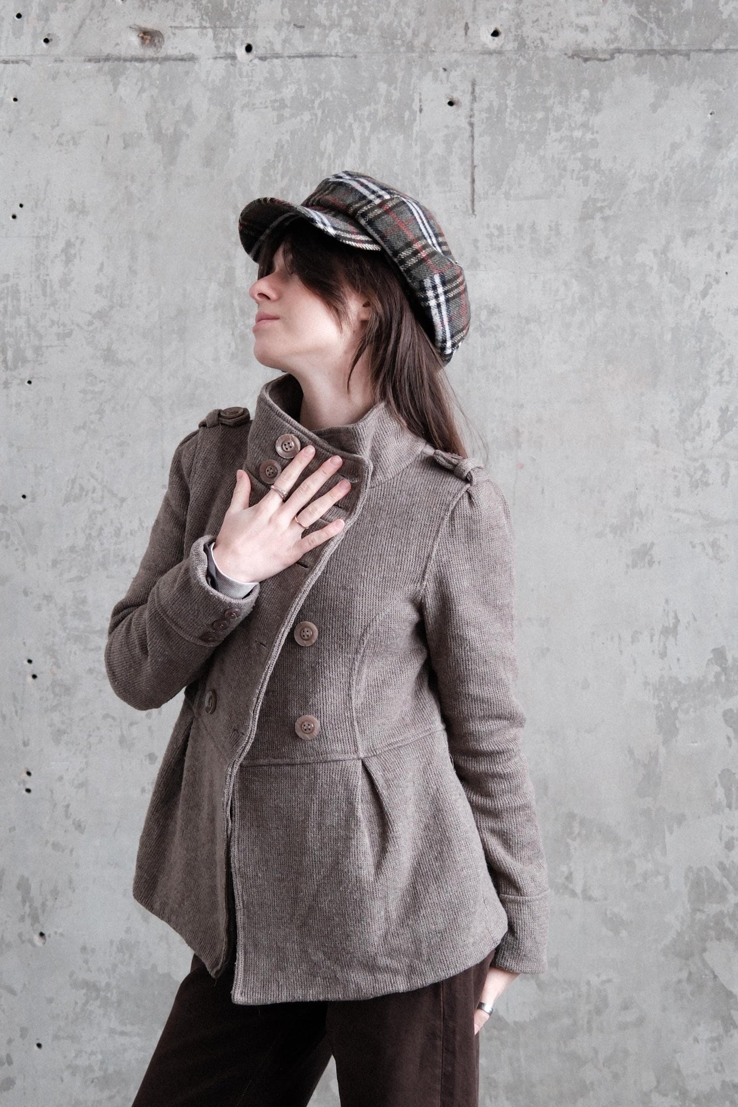 A person wearing a brown coat and plaid hat is lying on their back, with their head tilted back and hands covering their mouth.
