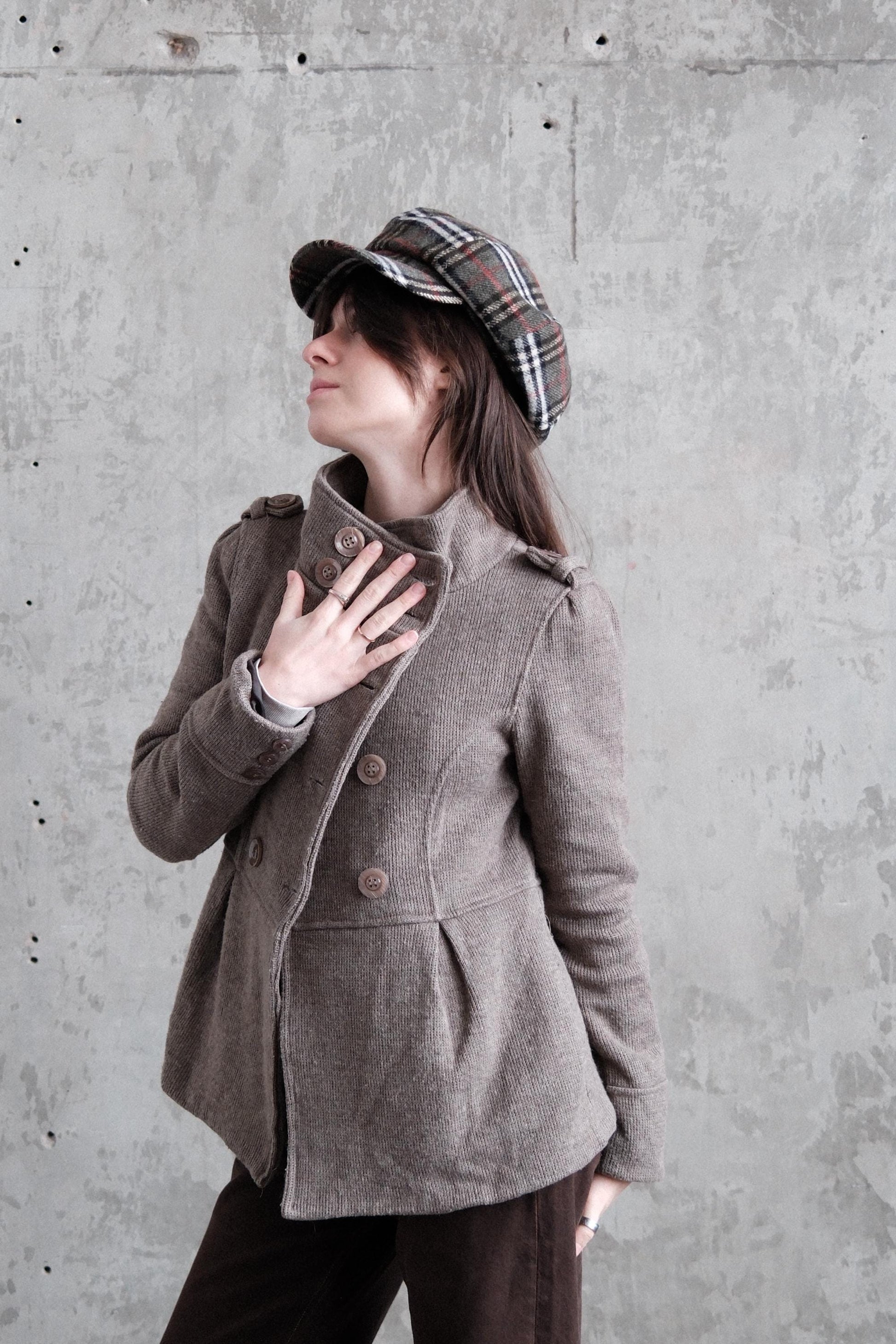 A person wearing a brown coat and plaid hat is lying on their back, with their head tilted back and hands covering their mouth.