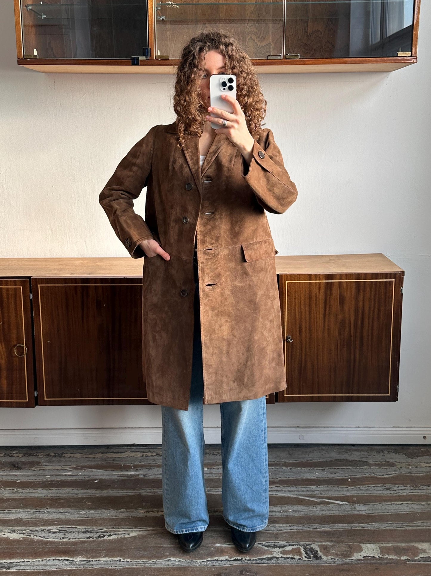 A person with curly hair is taking a selfie with a white phone, wearing a brown coat and blue jeans.