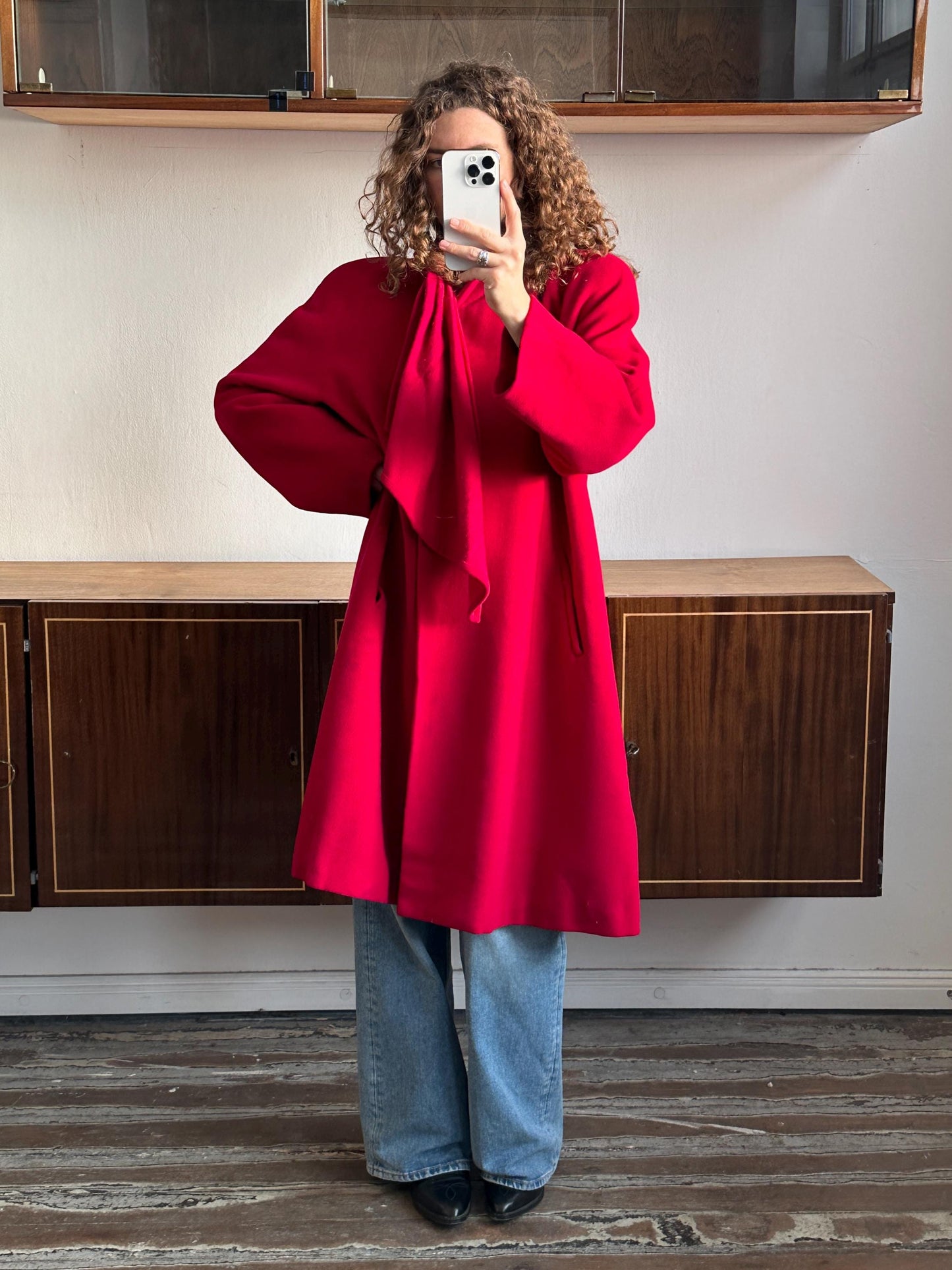A person wearing a red coat is taking a selfie with a smartphone in front of a wooden cabinet.