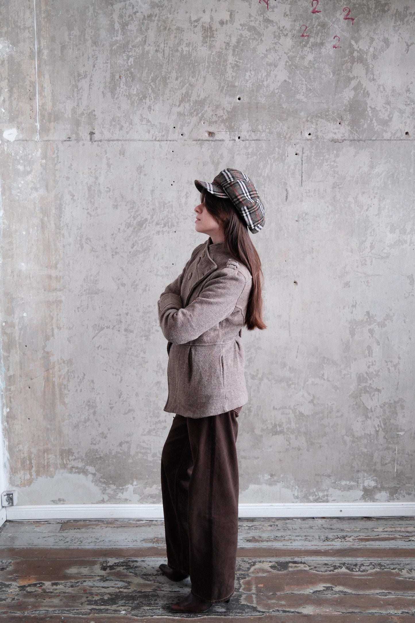 A person wearing a brown coat and plaid hat stands in a room with a concrete wall.