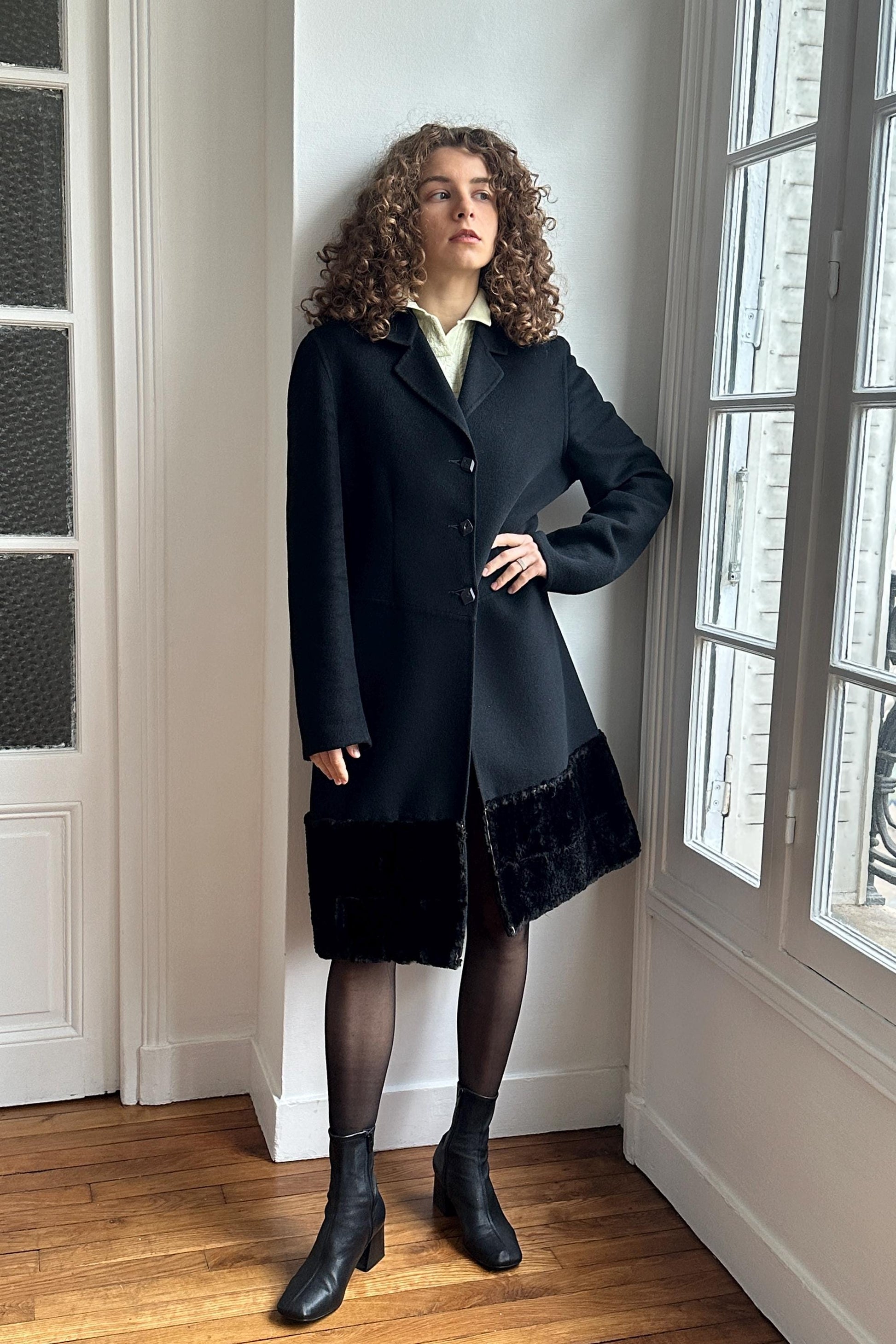 A woman in a black coat with fur trim stands in a room with a window, looking off to the side.