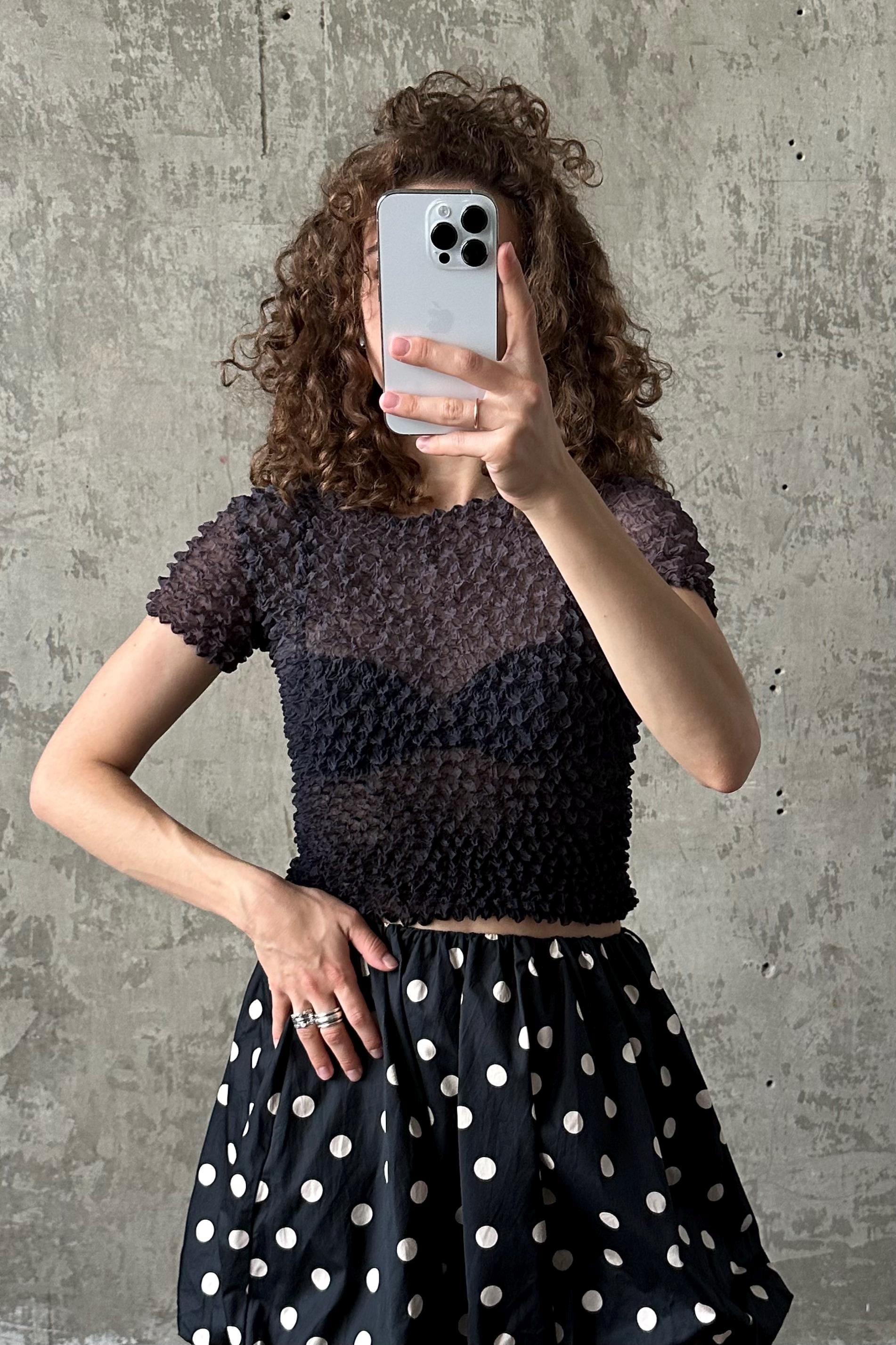 A woman with curly hair is taking a selfie with her iPhone, wearing a black and white polka dot skirt and a black top with a ruffled collar.