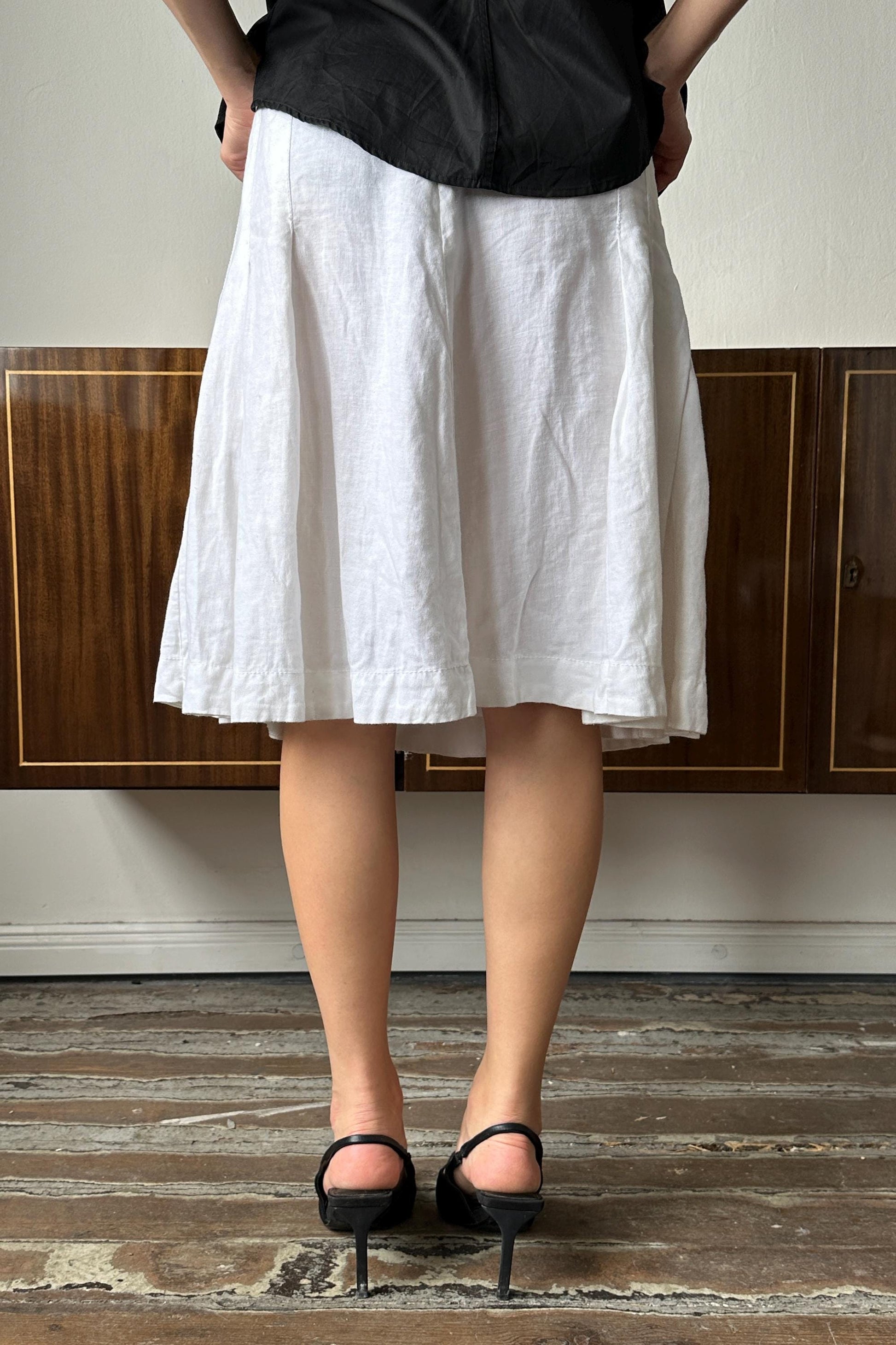 A person wearing a white skirt and black heels stands in front of a wooden cabinet.
