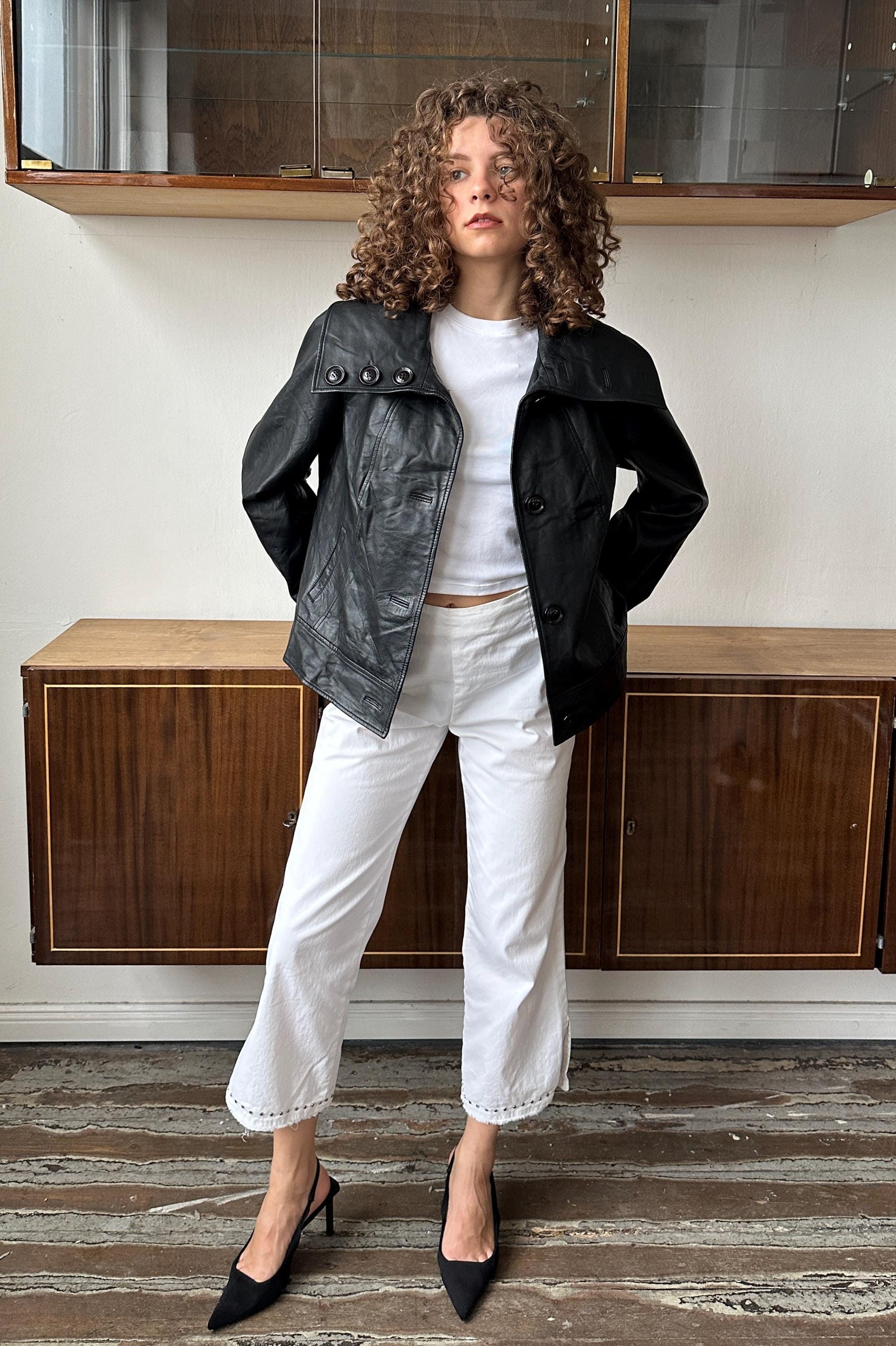 A woman with curly hair wearing a black leather jacket, white pants, and black heels stands in a room with wooden cabinets.