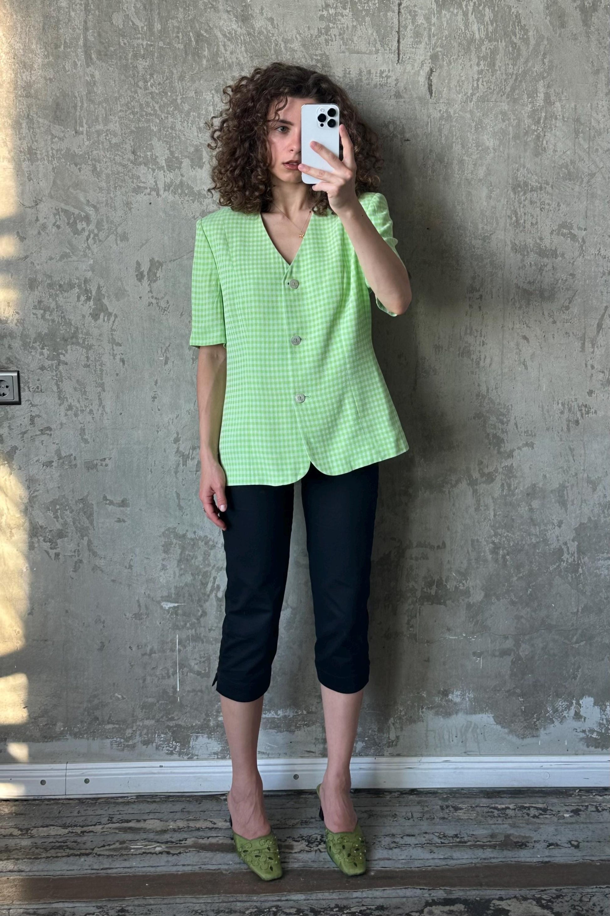 A woman with curly hair is taking a selfie with a white phone, wearing a green and white checkered blazer, black pants, and green heels.