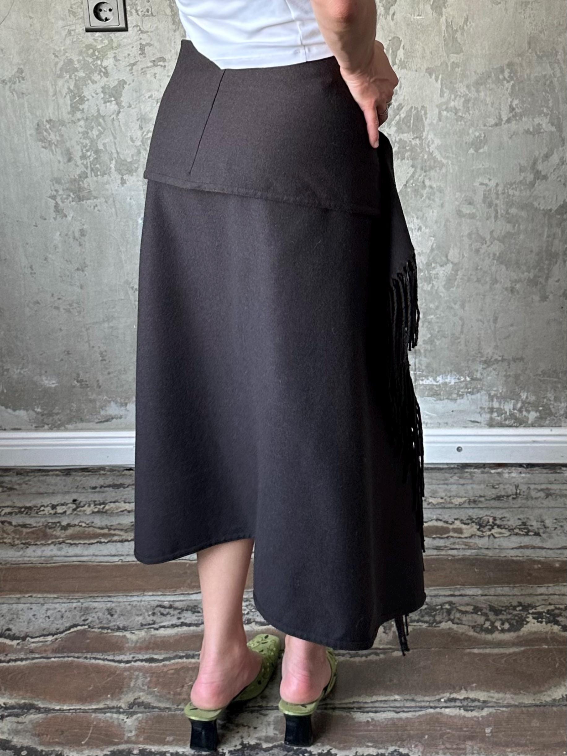 A person wearing a black skirt with fringe details, standing in front of a concrete wall.