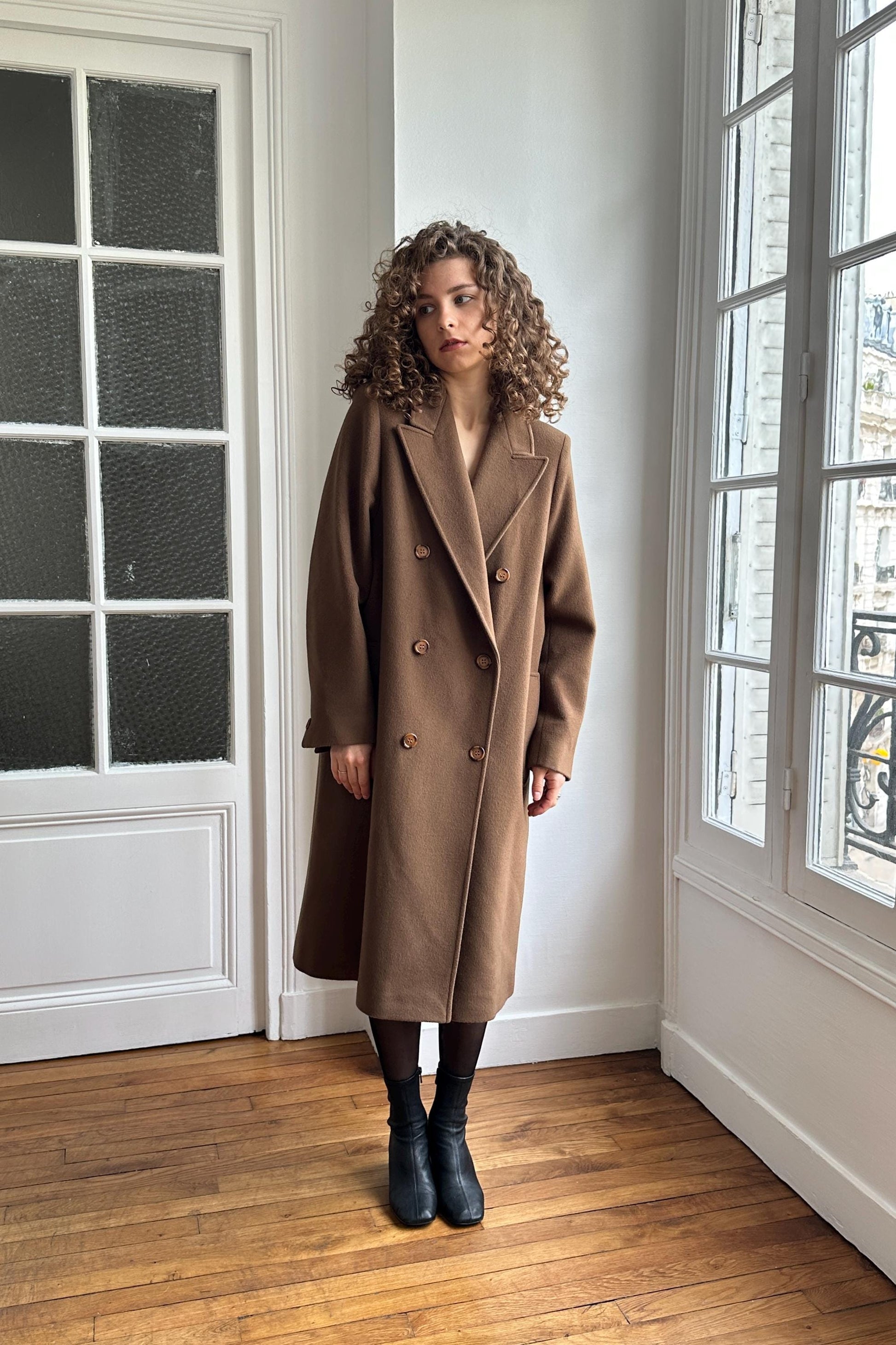 A woman in a brown coat stands in a room with large windows, looking off to the side.