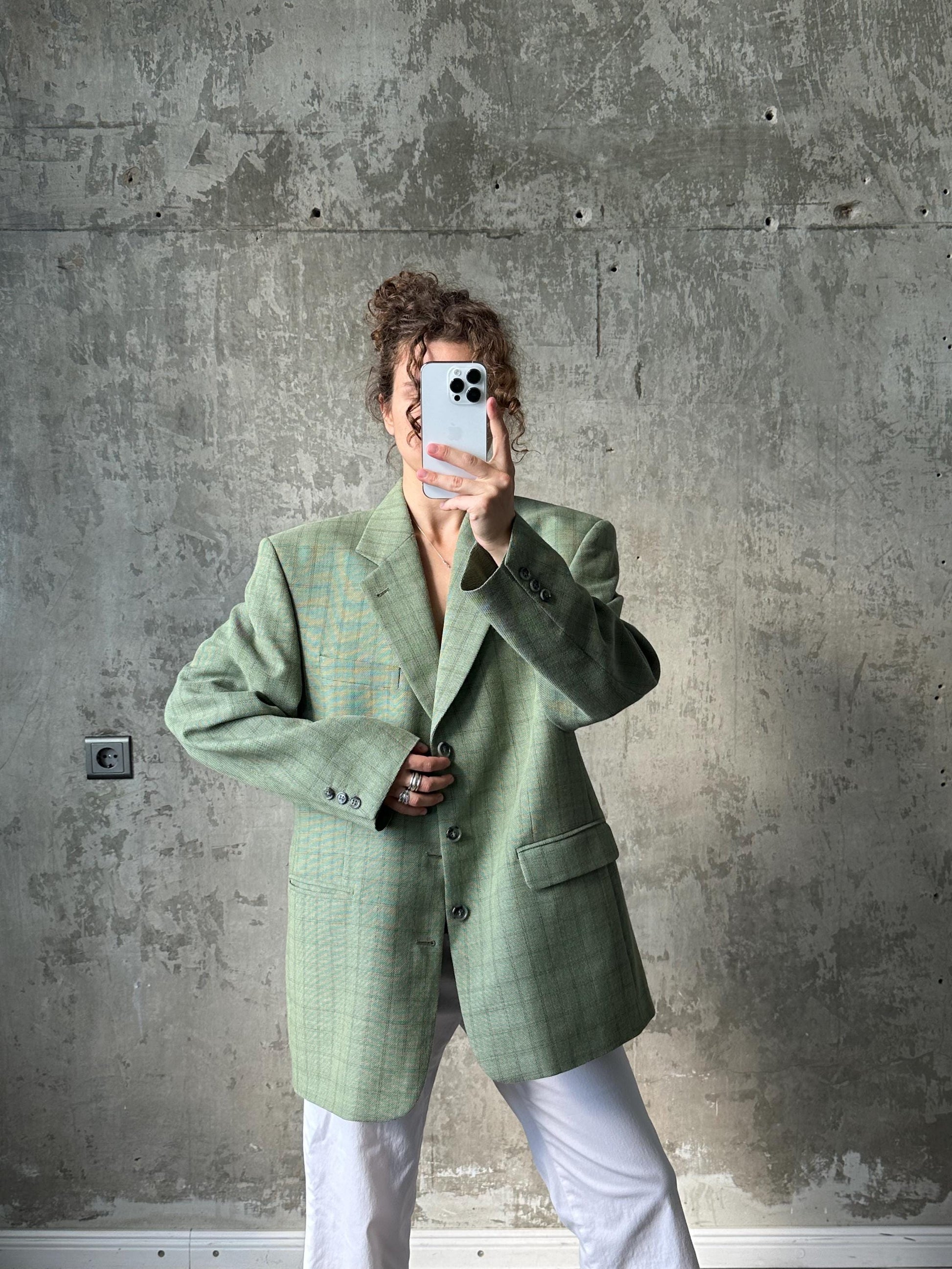 A person is taking a selfie with a white phone, wearing a green plaid jacket and white pants, against a concrete wall background.