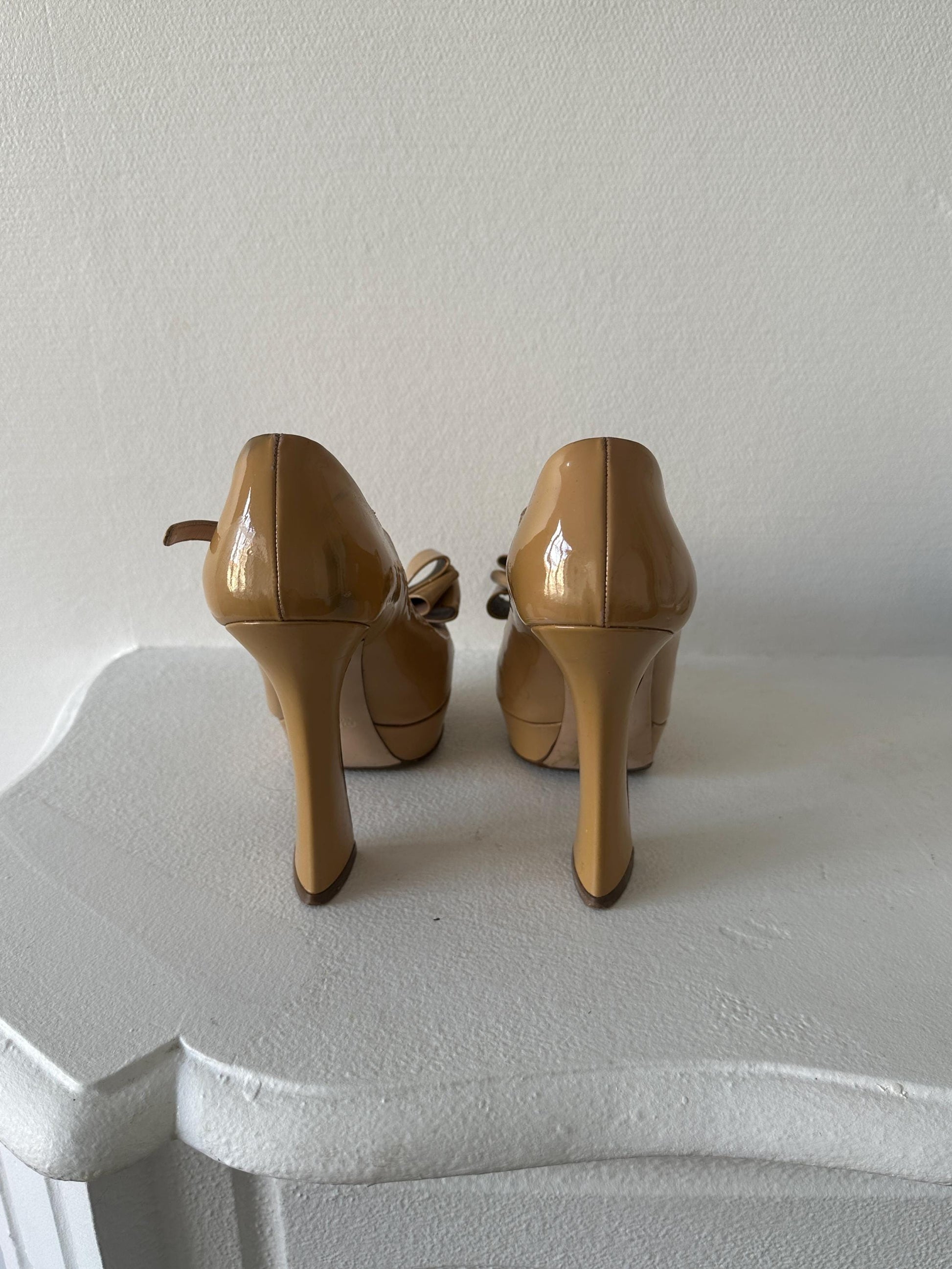 A pair of tan high-heeled shoes with a bow detail on the toe, placed on a white surface.
