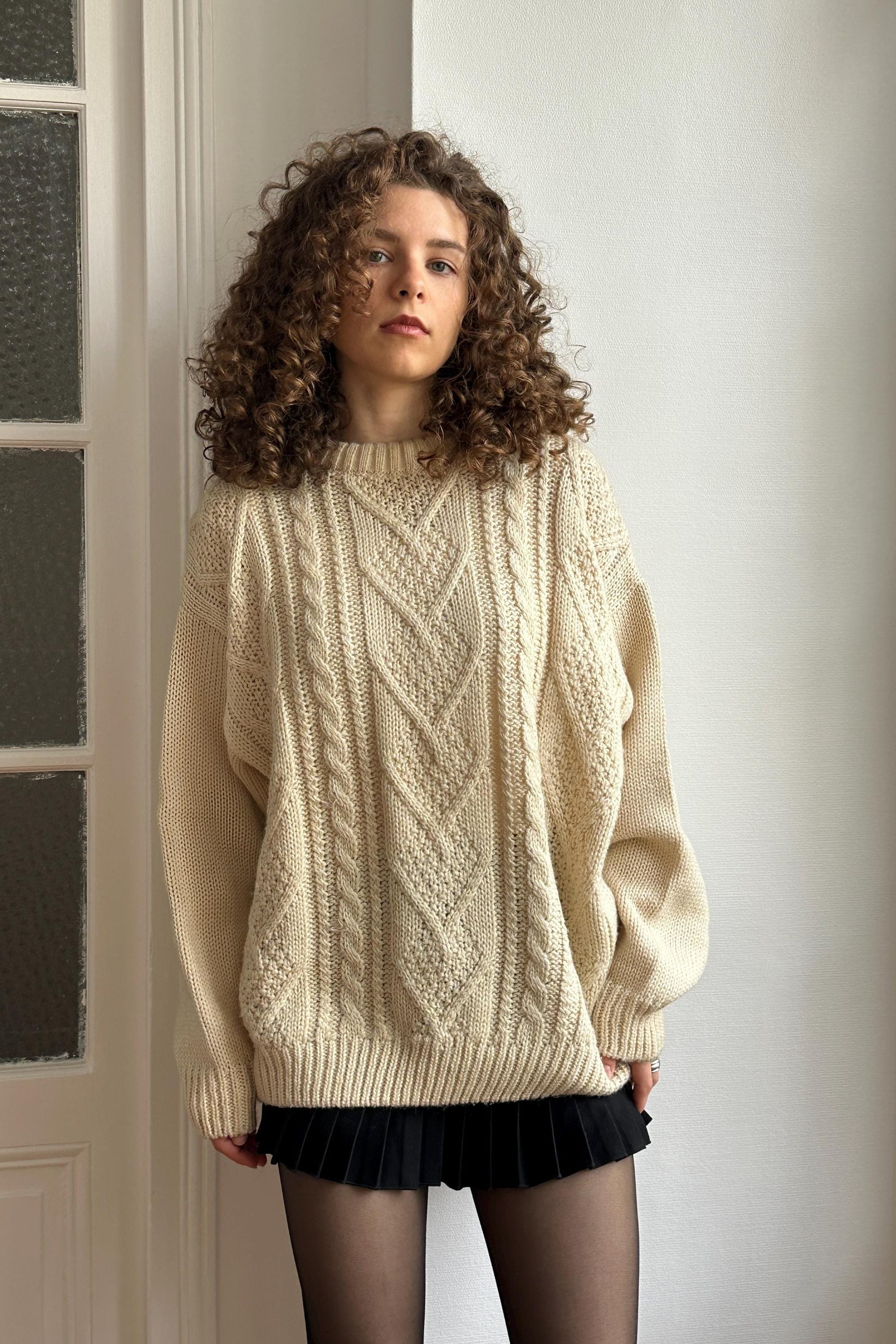 A young woman with curly hair stands in front of a white wall, wearing a cream-colored sweater with a geometric pattern and black shorts.